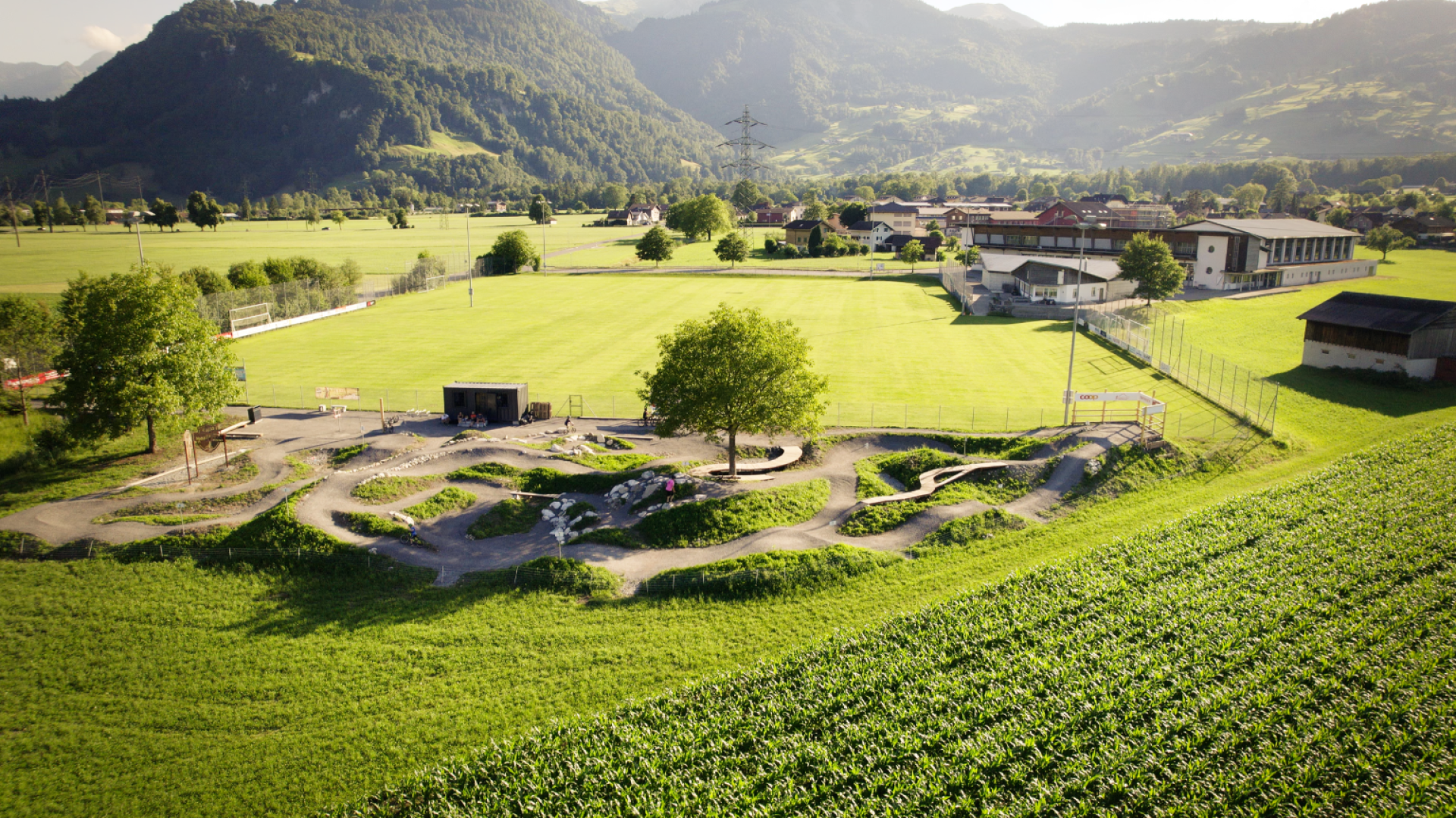 Luftaufnahme einer großen grünen Wiese, die von Bergen umgeben ist, mit einem gepflasterten Fahrrad- oder Skatepark, Bäumen und Gebäuden in der Nähe, umgeben von Ackerland und ländlicher Landschaft.