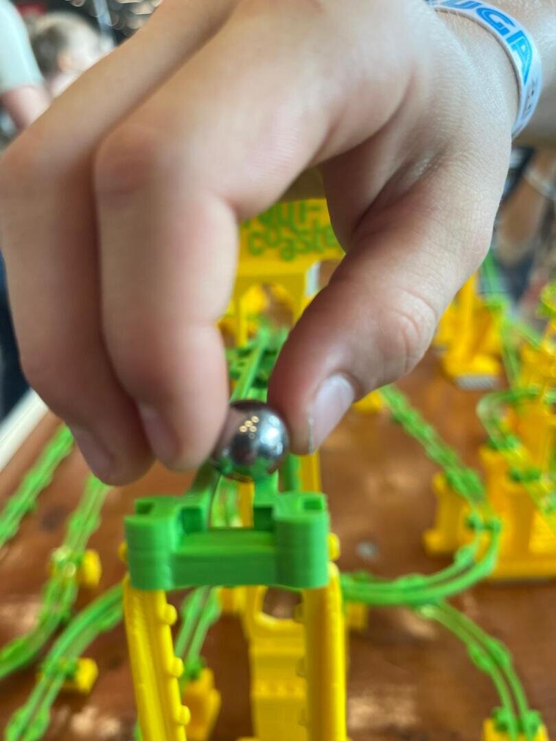 Nahaufnahme einer Hand, die eine Metallmurmel oben auf eine bunte Spielzeug-Achterbahn mit grünen Schienen und gelben Stützen legt. Der Hintergrund ist unscharf.