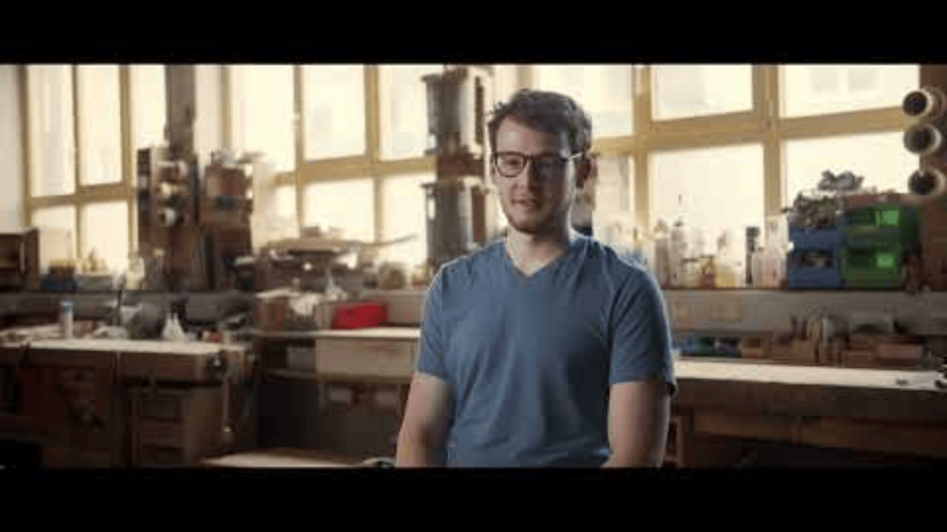 Ein Mann mit Brille und blauem T-Shirt sitzt lächelnd in einer Werkstatt, hinter ihm sind Werkzeuge, Geräte und Regale zu sehen, die von großen Fenstern beleuchtet werden.