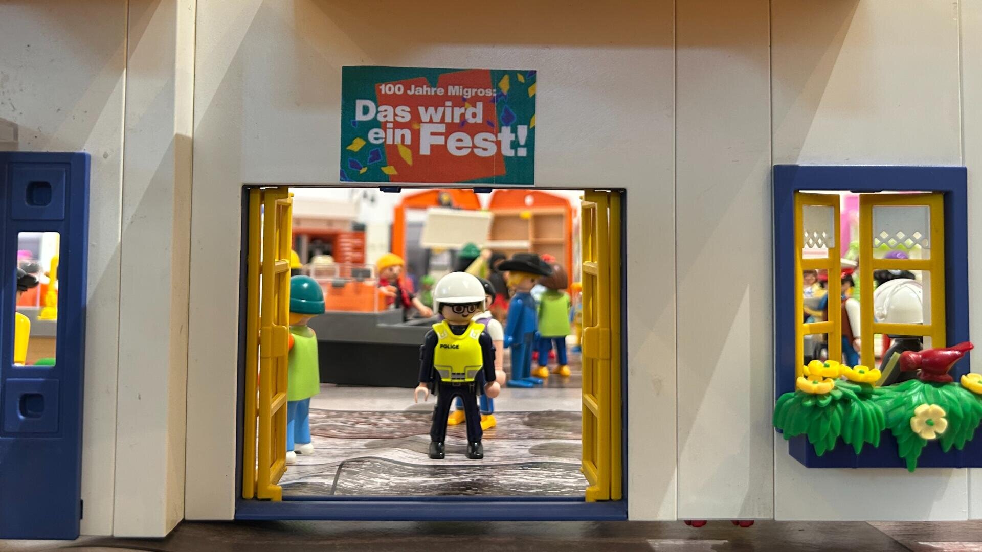 Eine Playmobil-Szene zeigt eine Figur in Bauweste und Helm, die in einer Tür unter einem Schild mit der Aufschrift Das wird ein Fest! Im Inneren des Gebäudes sind bunte Spielzeugleute und Dekorationen zu sehen.