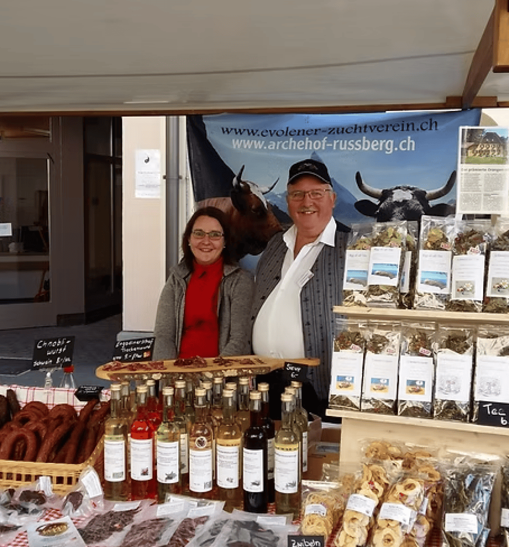 Ein Mann und eine Frau stehen hinter einem Marktstand, an dem verschiedene verpackte Lebensmittel, Würste und Getränkeflaschen angeboten werden. Hinter ihnen hängt ein Transparent mit Kühen und Internetadressen. Beide lächeln und sind warm angezogen.