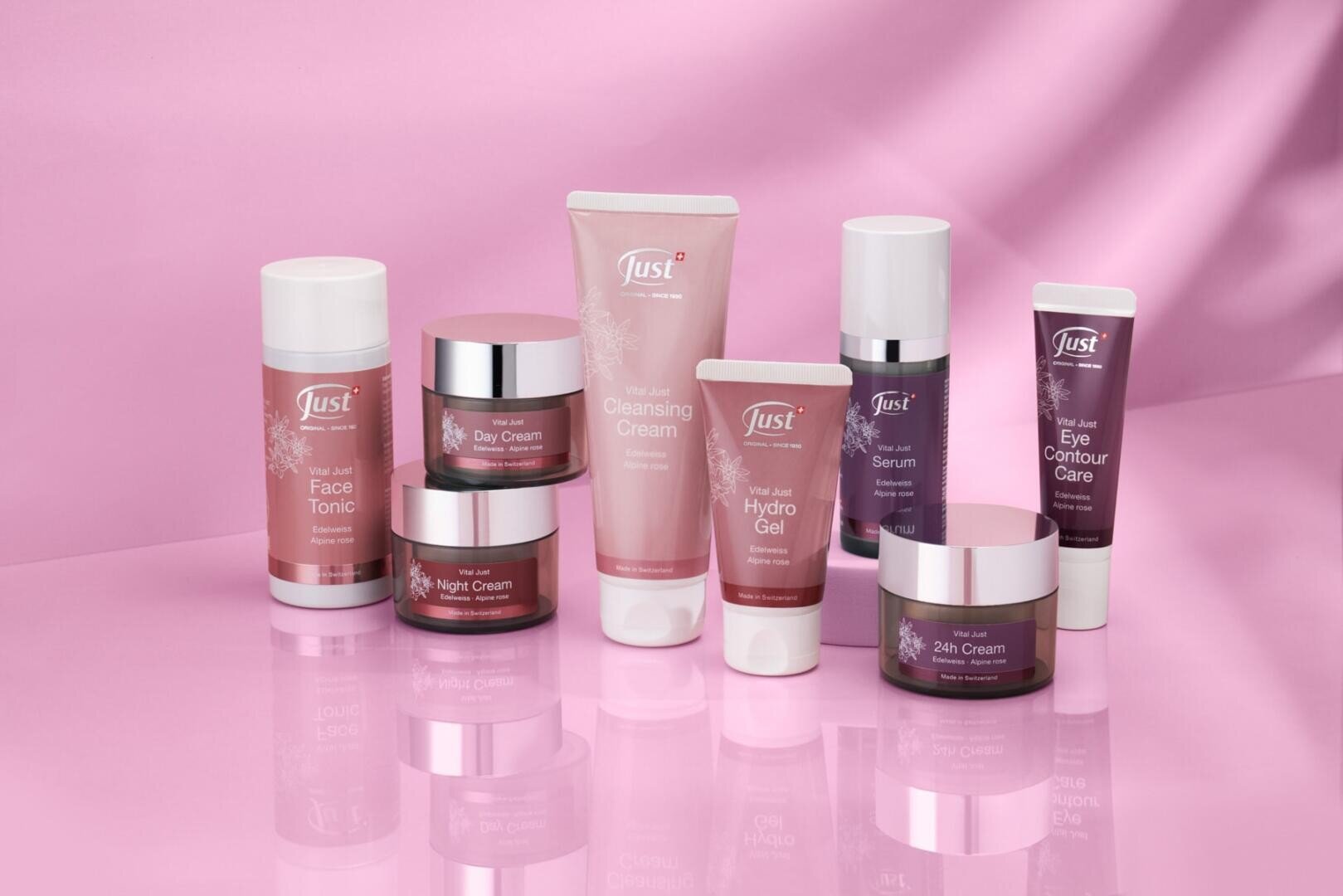 Ein Set mit neun rosafarbenen und weißen Hautpflegeprodukten, darunter Cremes, Gels und Seren, die ordentlich auf einer glänzenden rosafarbenen Oberfläche mit einem zartrosa Hintergrund angeordnet sind.