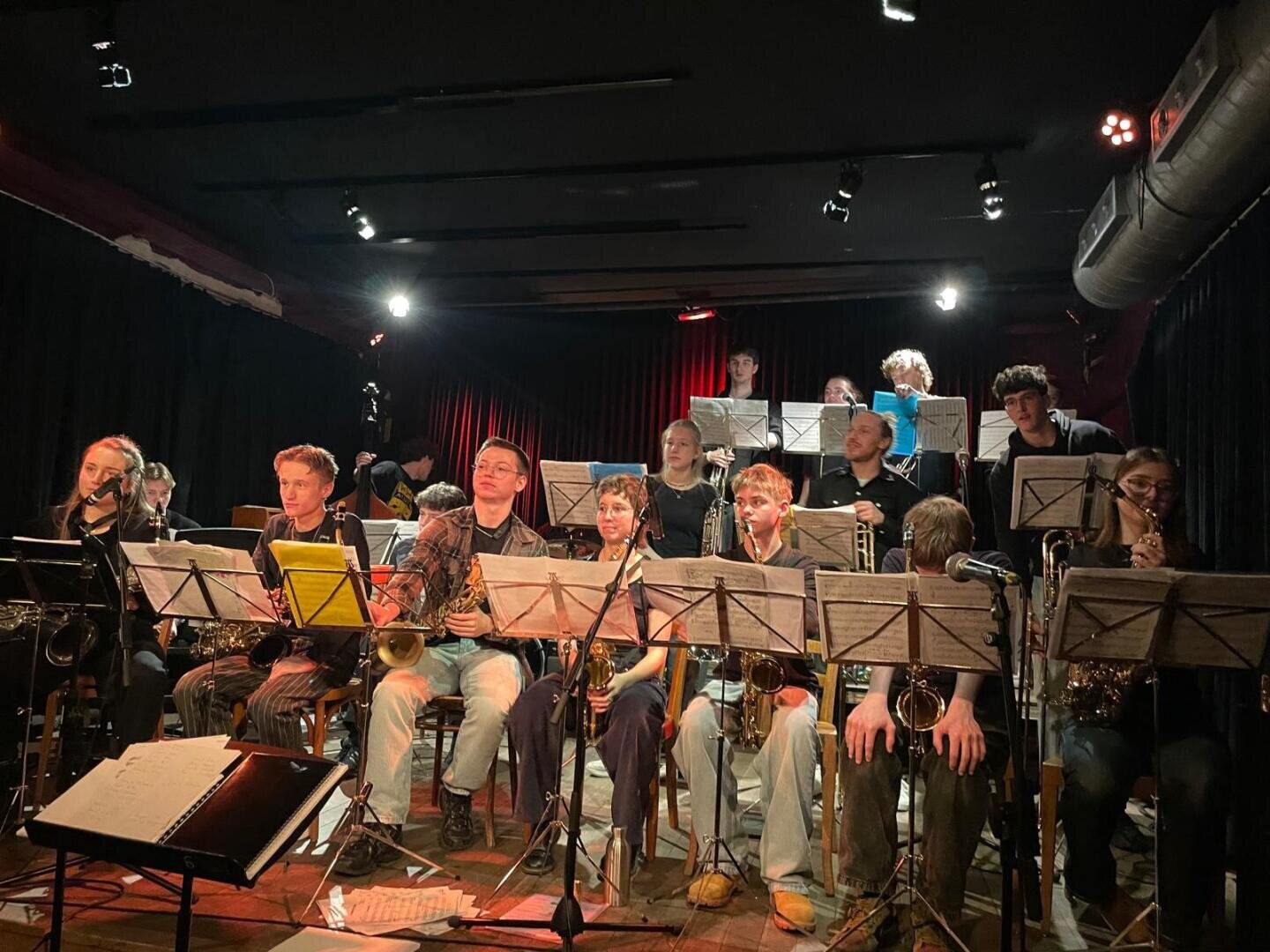Auf der Bühne spielt eine etwa 17-köpfige Jazzband mit verschiedenen Blech- und Holzblasinstrumenten, in Reihen sitzend, mit Notenständern davor, unter warmer Bühnenbeleuchtung und vor dunklem Hintergrund.