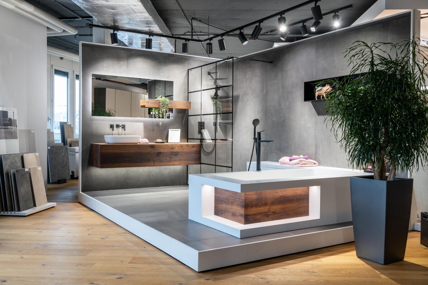 Ein moderner Badezimmer-Showroom verfügt über eine große Badewanne mit Holzakzenten, einen schwebenden Waschtisch, wandmontierte Regale, eine Glasdusche, Topfpflanzen und eine sanfte Beleuchtung, alles auf einer erhöhten Plattform mit grauem und Holzfußboden.