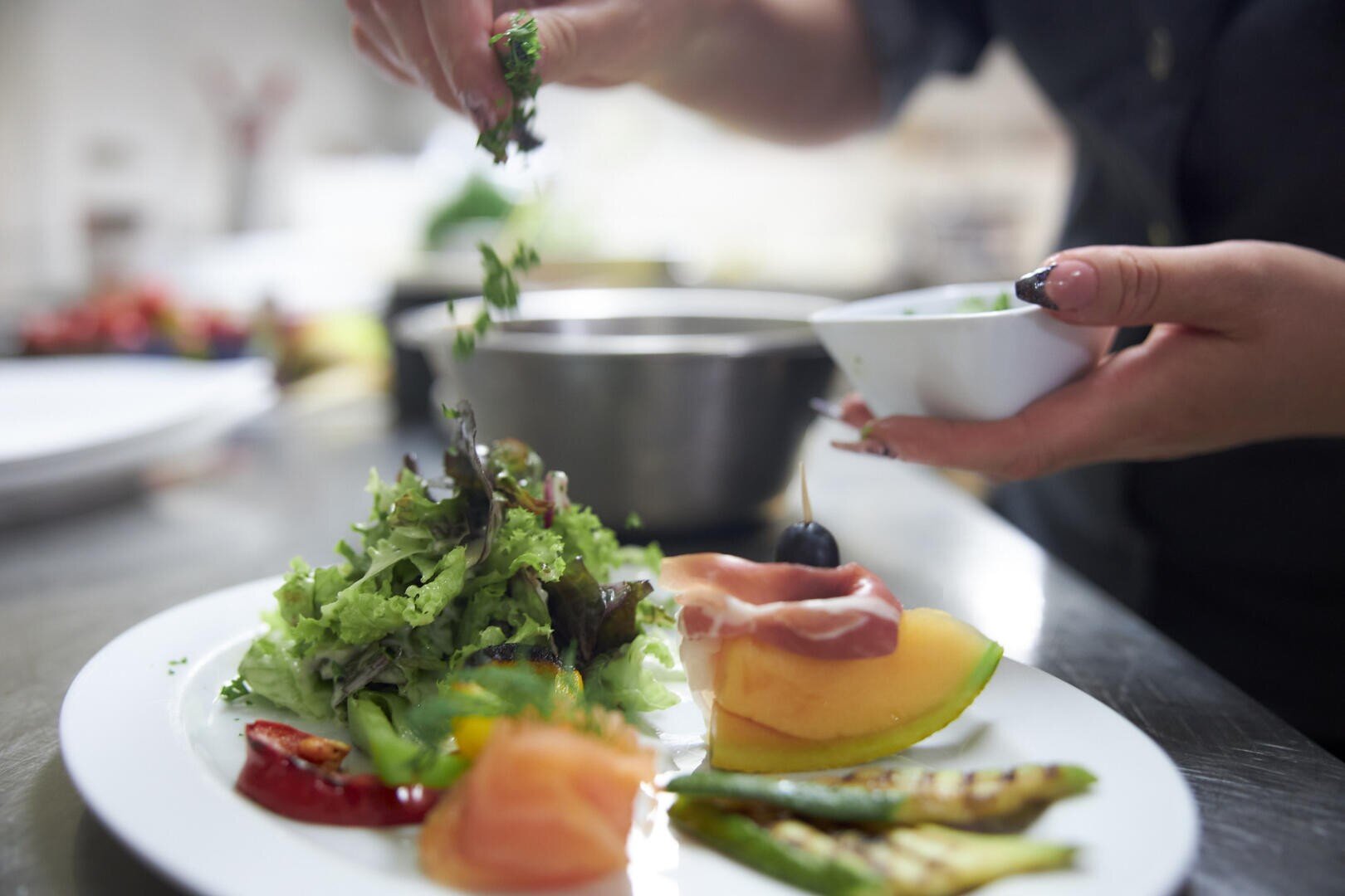 Ein Koch garniert einen Teller mit Salat und verschiedenen frischen Gemüsesorten mit gehackten Kräutern und verleiht dem farbenfrohen Gericht auf einem Küchentisch aus Edelstahl den letzten Schliff.