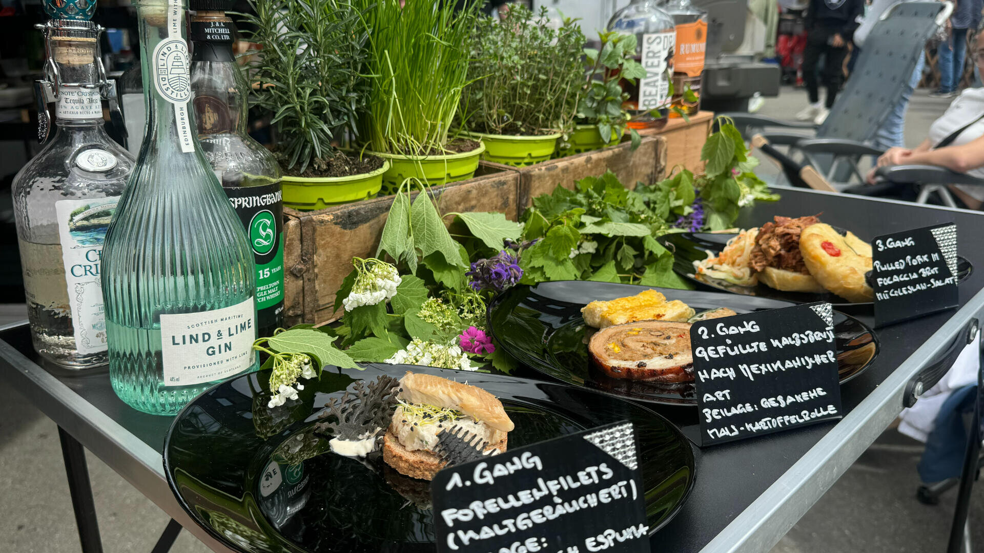 Drei schwarze Teller mit Gourmetgerichten und handgeschriebenen Etiketten stehen auf einem Tisch mit frischen Kräutern, Blattgemüse und essbaren Blumen. Dahinter stehen Flaschen mit Gin und anderen Spirituosen sowie Kräuter und Pflanzen in Töpfen.