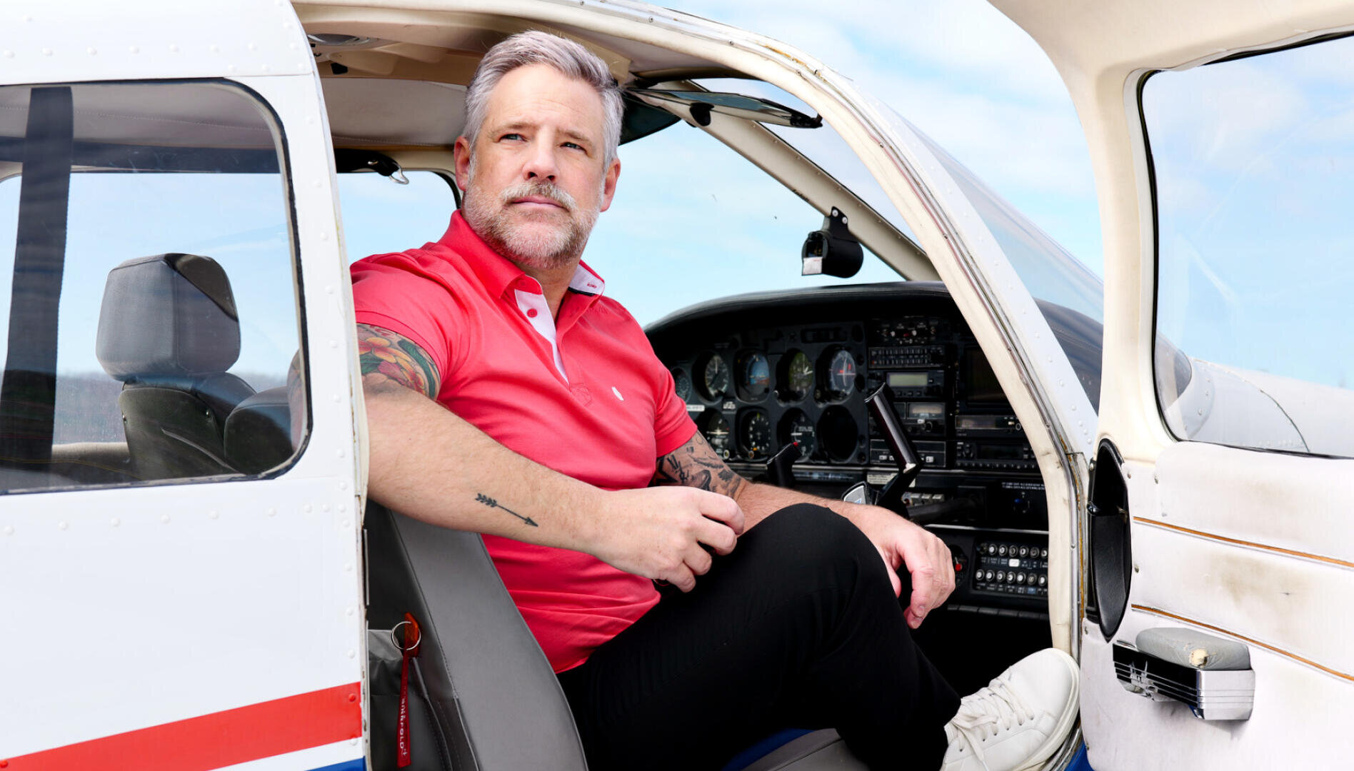 Ein Mann mit grauem Haar und Bart, der ein rotes Polohemd und eine schwarze Hose trägt, sitzt in der offenen Tür eines Kleinflugzeugs und schaut nachdenklich in die Ferne. Hinter ihm sind die Instrumente im Cockpit zu sehen.