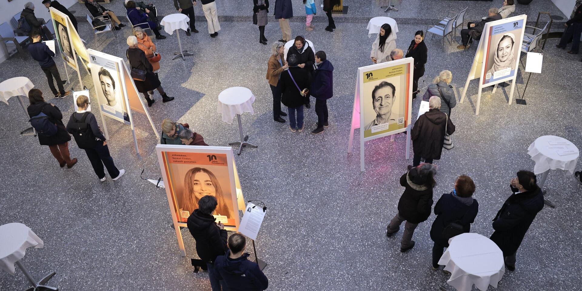 Die Menschen gehen umher und betrachten große Porträts in einer Innenausstellung. Kleine runde Tische sind im Raum verstreut, und Gruppen und Einzelpersonen betrachten die Kunstwerke und unterhalten sich. Die Umgebung wirkt hell und großzügig.