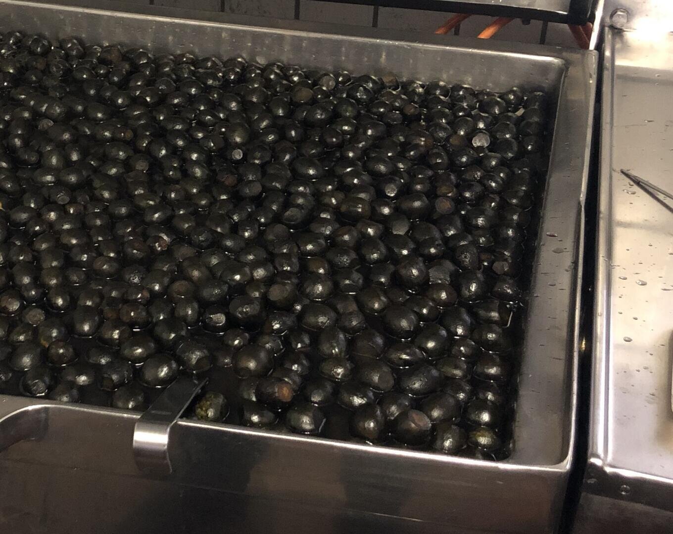 Ein großer Behälter aus rostfreiem Stahl, gefüllt mit vielen dunklen, glänzenden schwarzen Wasserkastanien, die teilweise in Wasser getaucht sind, in einem Raum, der wie eine Großküche oder ein Lebensmittelverarbeitungsraum aussieht.
