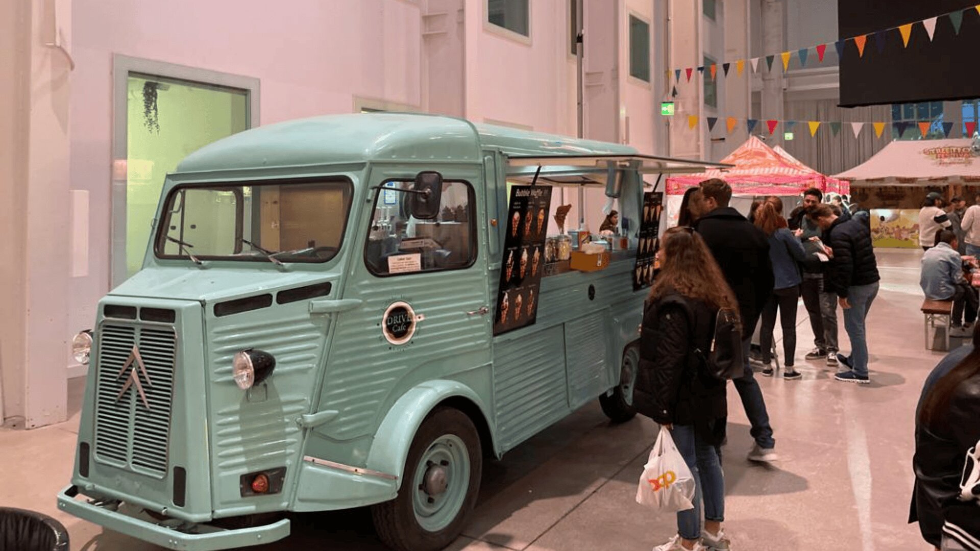 Ein alter hellblauer Imbisswagen ist drinnen geparkt. Mehrere Menschen stehen in der Schlange, um Essen zu bestellen. Über dem Wagen hängen bunte Fahnen, und im Hintergrund sind weitere Stände zu sehen. Die Umgebung sieht festlich und geschäftig aus.