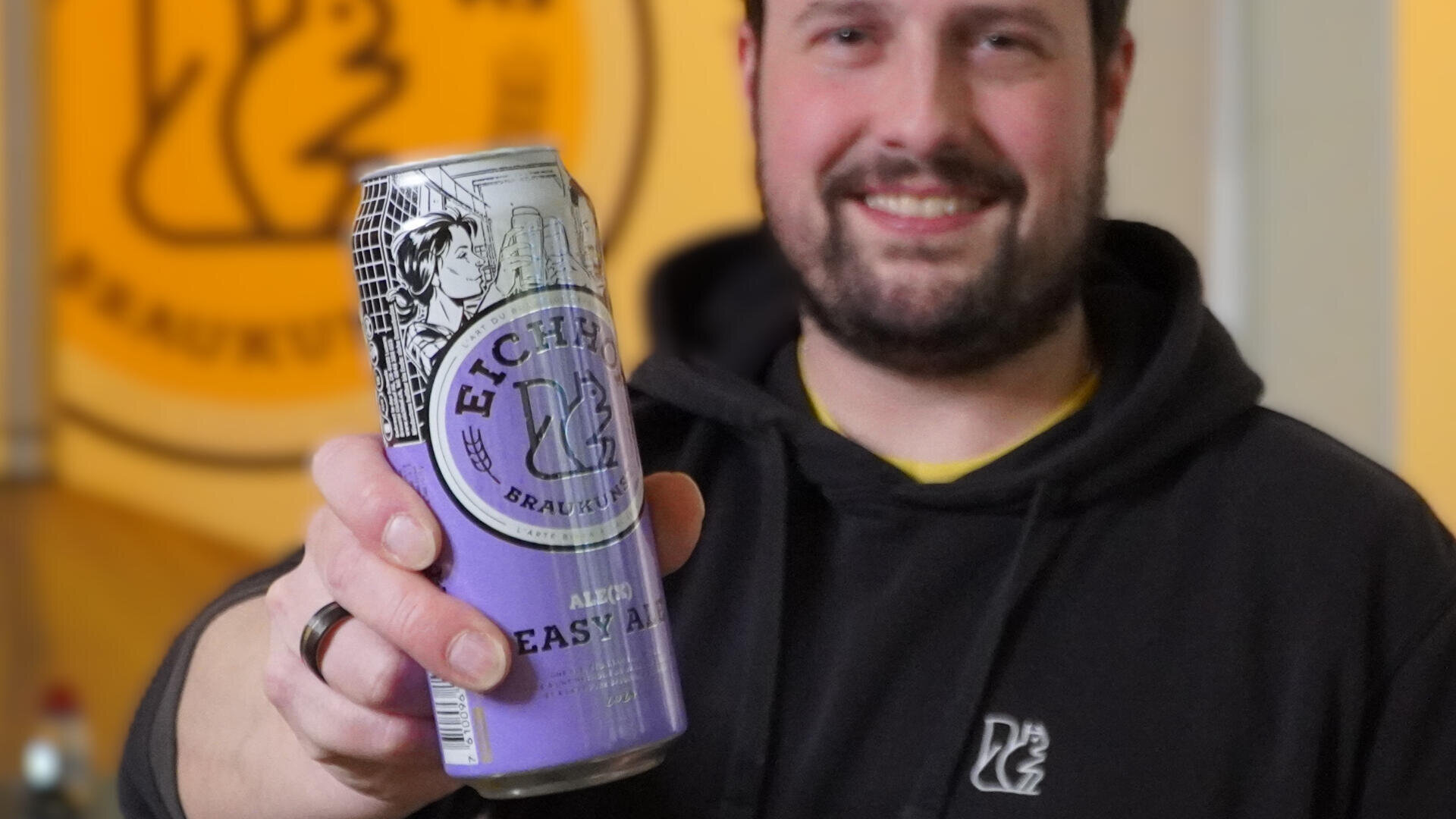 Eine Person in einem schwarzen Kapuzenpulli mit Logo lächelt und hält eine Dose Eichbaum Easy Ale Bier in die Kamera, mit einem verschwommenen gelben Logo im Hintergrund.