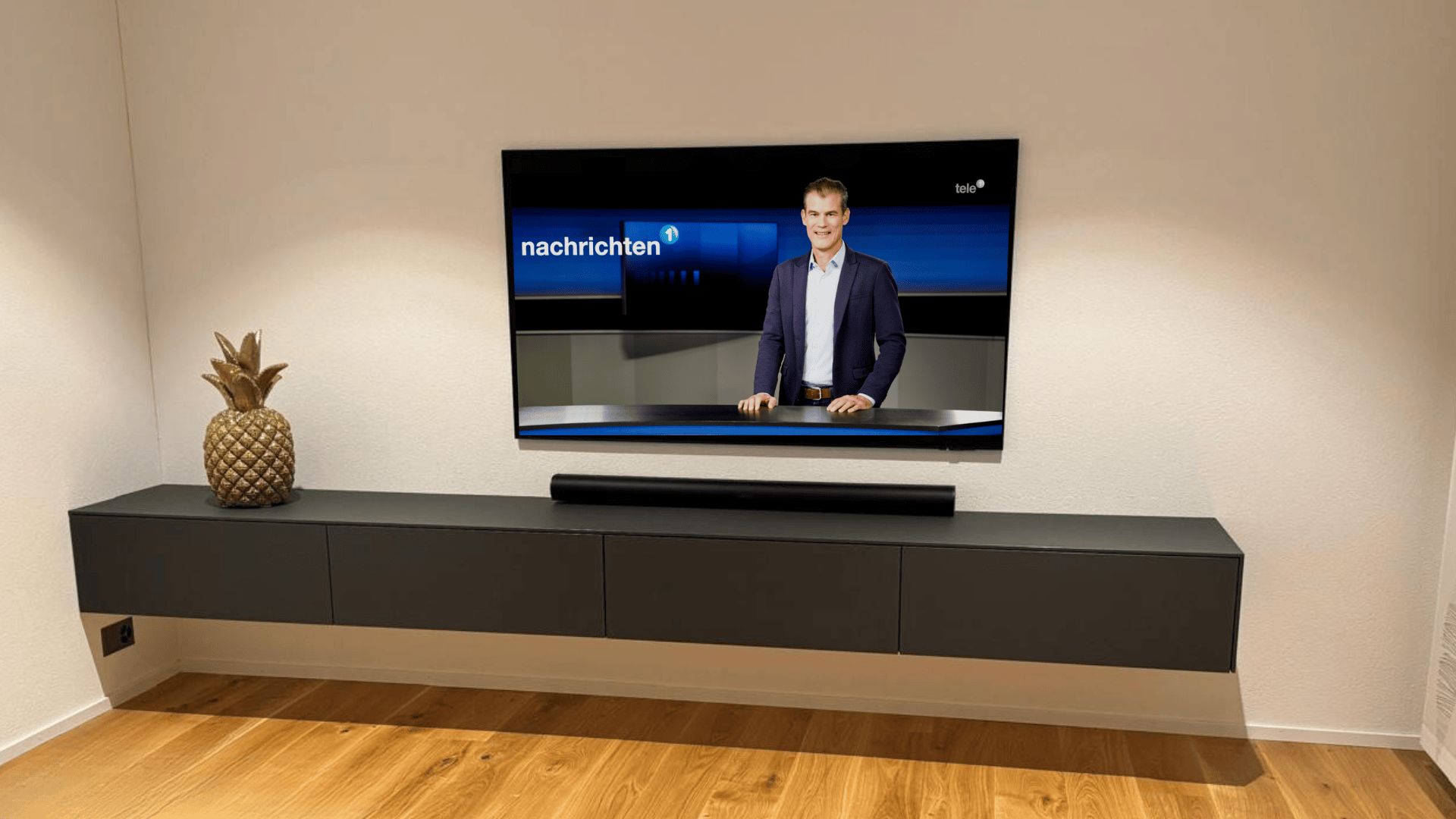 Ein modernes Wohnzimmer mit einem an der Wand montierten Fernseher, auf dem eine Nachrichtensendung mit einem männlichen Moderator zu sehen ist. Unter dem Fernseher steht ein schlichter schwarzer Schrank mit einer dekorativen Ananas auf der linken Seite. Der Raum hat einen Holzboden und weiße Wände.