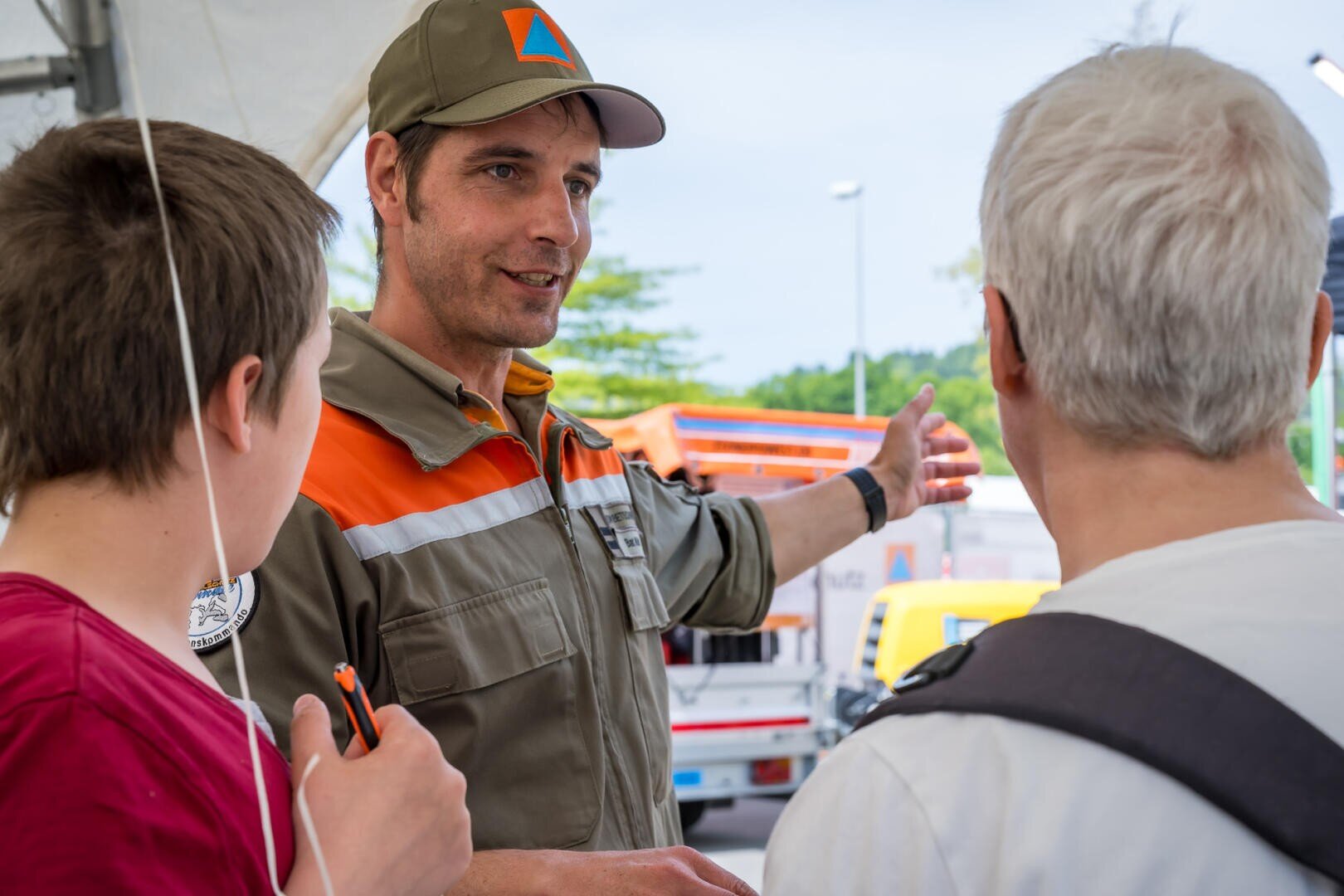 Ein Mann in Uniform gestikuliert, während er mit zwei Personen, einer älteren und einer jüngeren, bei einer Veranstaltung im Freien spricht, während im Hintergrund Einsatzfahrzeuge zu sehen sind.