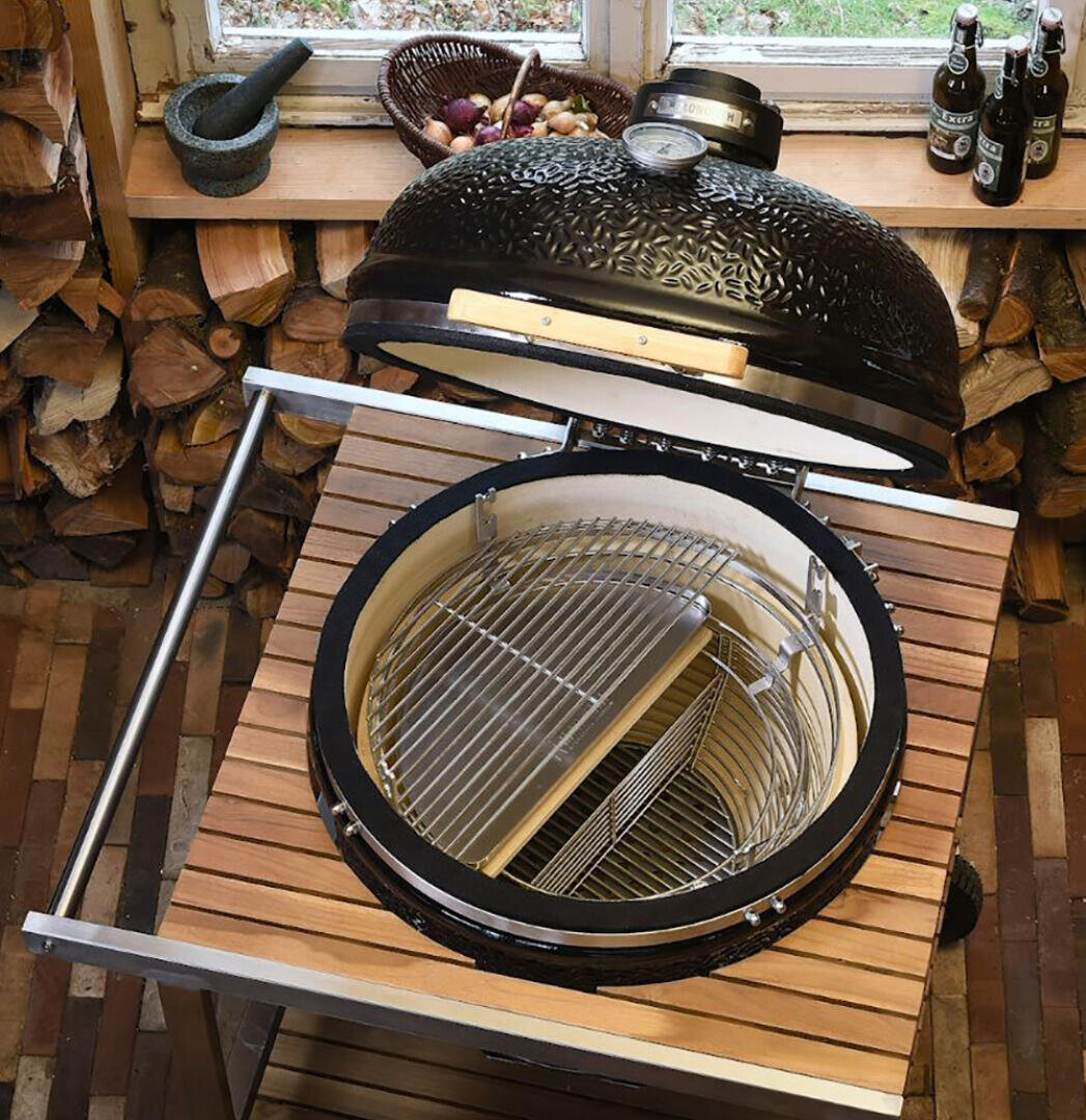 Eine Ansicht von oben auf einen offenen Keramikgrill mit Metallrosten, der auf einem Holztisch steht. Darunter ist Brennholz gestapelt; auf der Fensterbank stehen Flaschen und Zwiebeln. Der Deckel des Grills ist strukturiert und verfügt über ein Thermometer.
