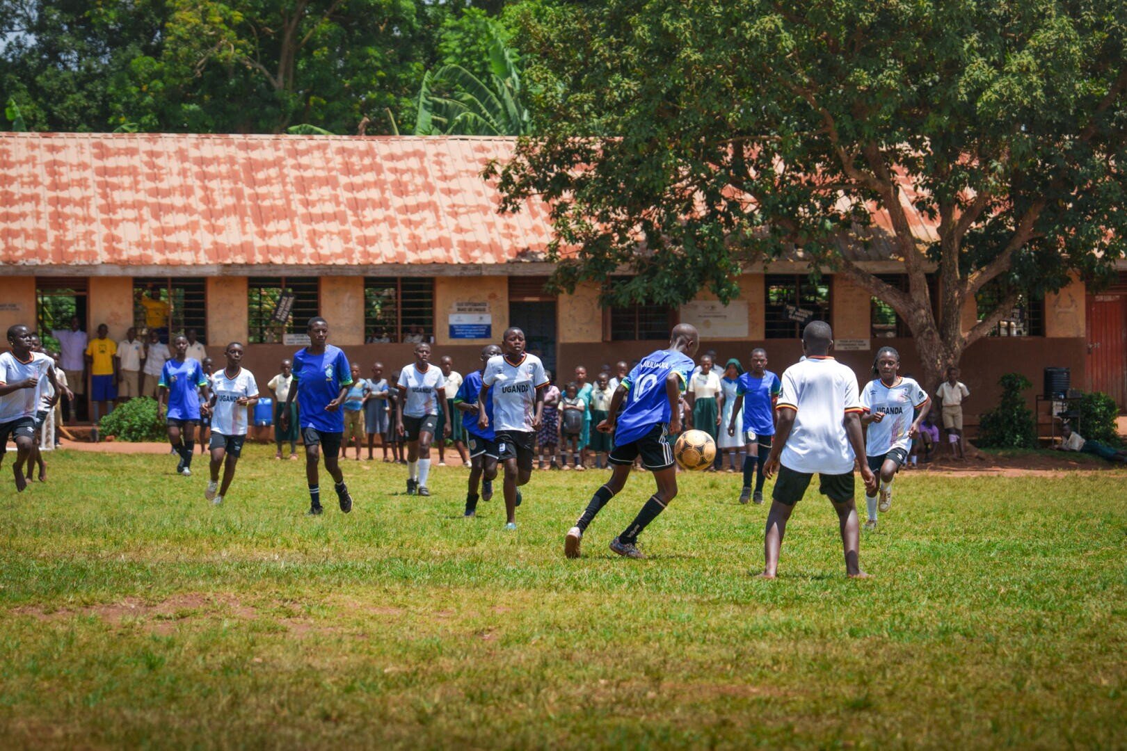 Kinder spielen Fußball auf einer Wiese, während eine Gruppe von Zuschauern vor einem rostfarbenen Gebäude und Bäumen im Hintergrund zusieht. Die meisten Spieler tragen blau-weiße Trikots.