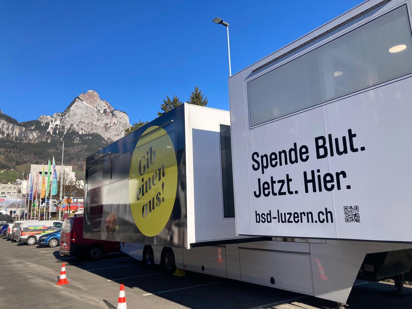 Ein mobiles Blutspendezentrum auf einem Parkplatz mit den Schweizer Alpen im Hintergrund. Auf den Schildern am Fahrzeug steht "Spende Blut. Jetzt. Hier." und "Gib einen aus." mit einem klaren blauen Himmel über sich.