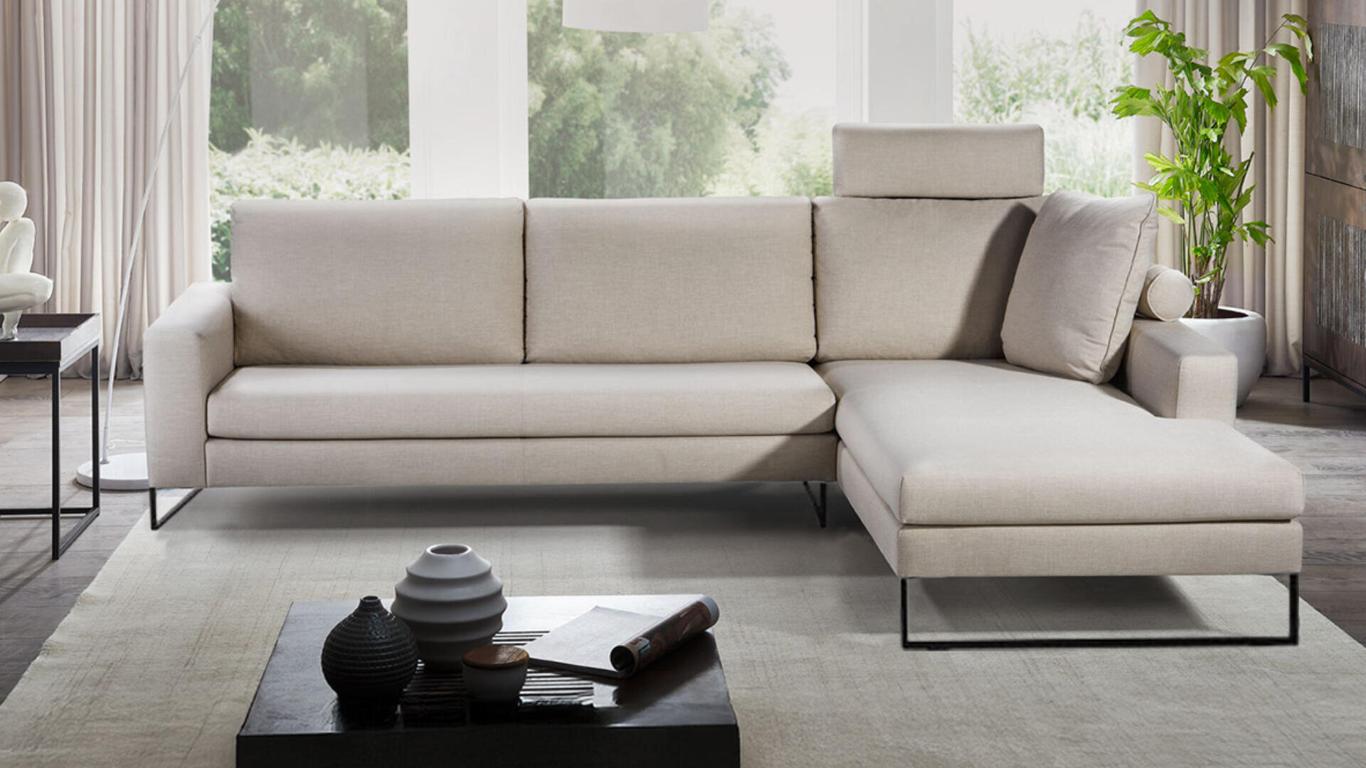 Ein modernes Wohnzimmer mit einem hellbeigen L-förmigen Sofa auf einem hellen Teppich, einem schwarzen Couchtisch mit dekorativen Vasen, Topfpflanzen und großen Fenstern, die natürliches Licht hereinlassen.