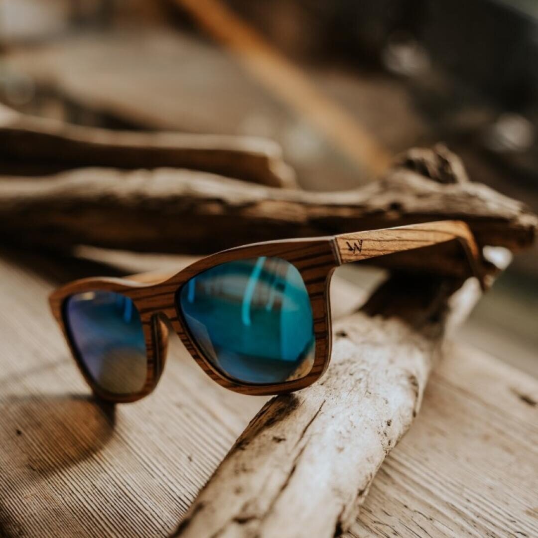 Die hölzerne Sonnenbrille mit blau verspiegelten Gläsern ruht auf Treibholzstücken auf einer rustikalen Holzoberfläche und schafft eine natürliche und entspannte Atmosphäre.