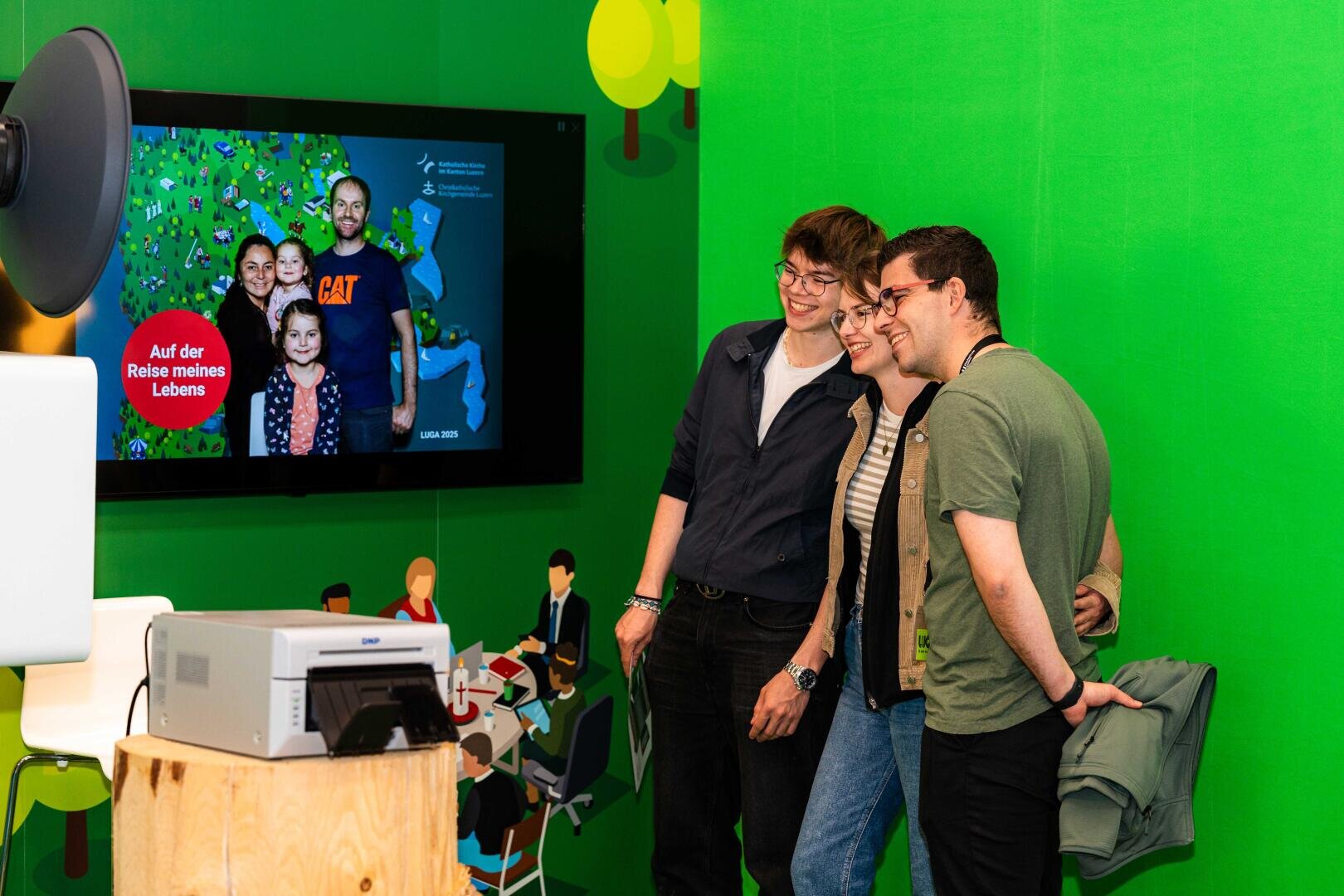 Vier Personen stehen dicht beieinander und lächeln für ein Foto vor einem grünen Hintergrund. Hinter ihnen zeigt ein digitaler Bildschirm ein weiteres Gruppenfoto mit deutschem Text, und in der Nähe sind verschiedene technische Geräte zu sehen.