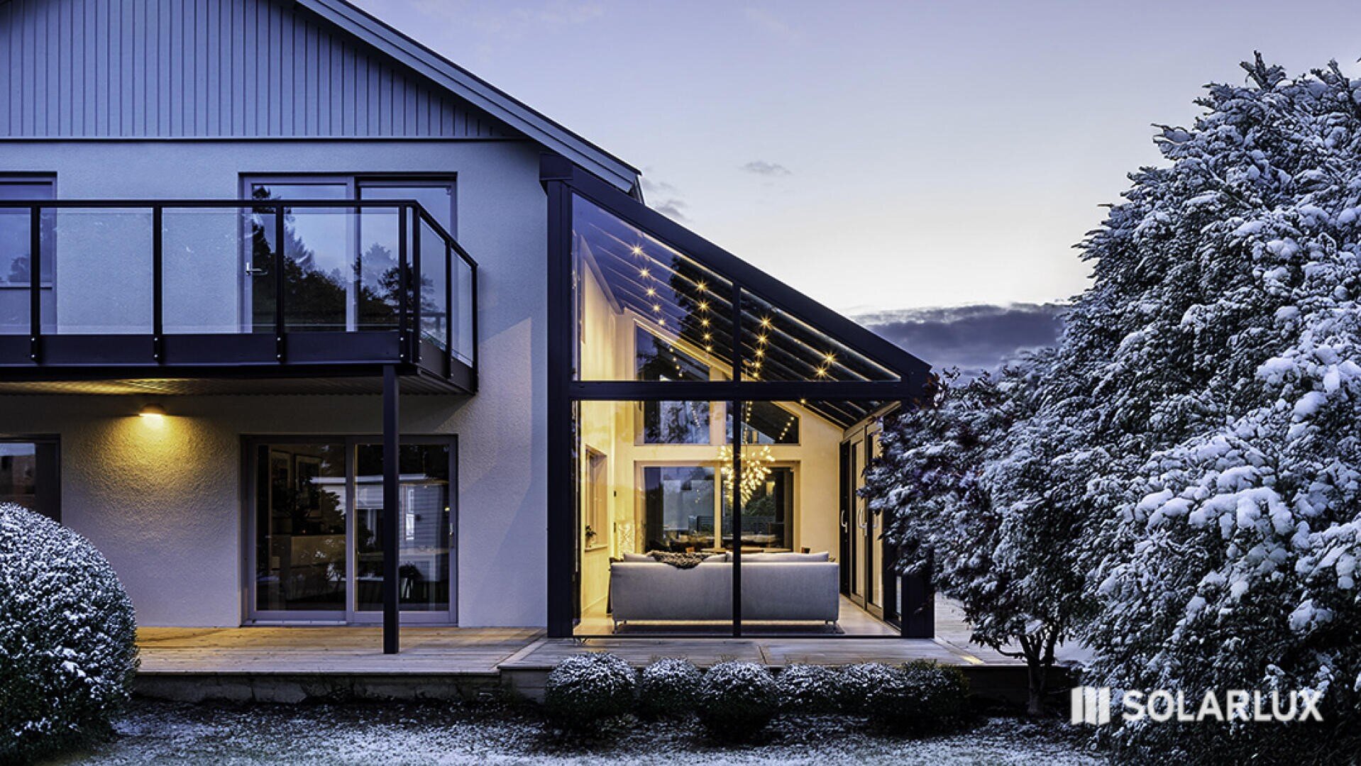 Modernes Haus mit großen Glasfenstern und einer überdachten Terrasse, umgeben von schneebedeckten Bäumen und Sträuchern in der Abenddämmerung. Der Innenraum ist warm beleuchtet, und das SOLARLUX-Logo ist in der rechten unteren Ecke zu sehen.