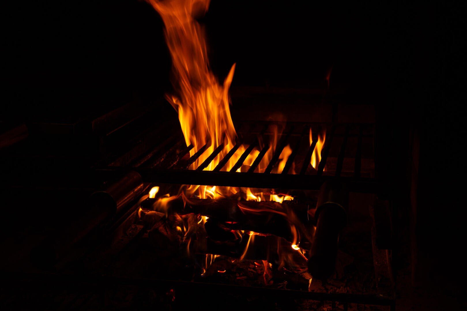 Ein Grill mit leuchtend orangefarbenen Flammen und glühender Glut vor einem dunklen Hintergrund, der an ein nächtliches Grillen im Freien oder ein Lagerfeuer erinnert.