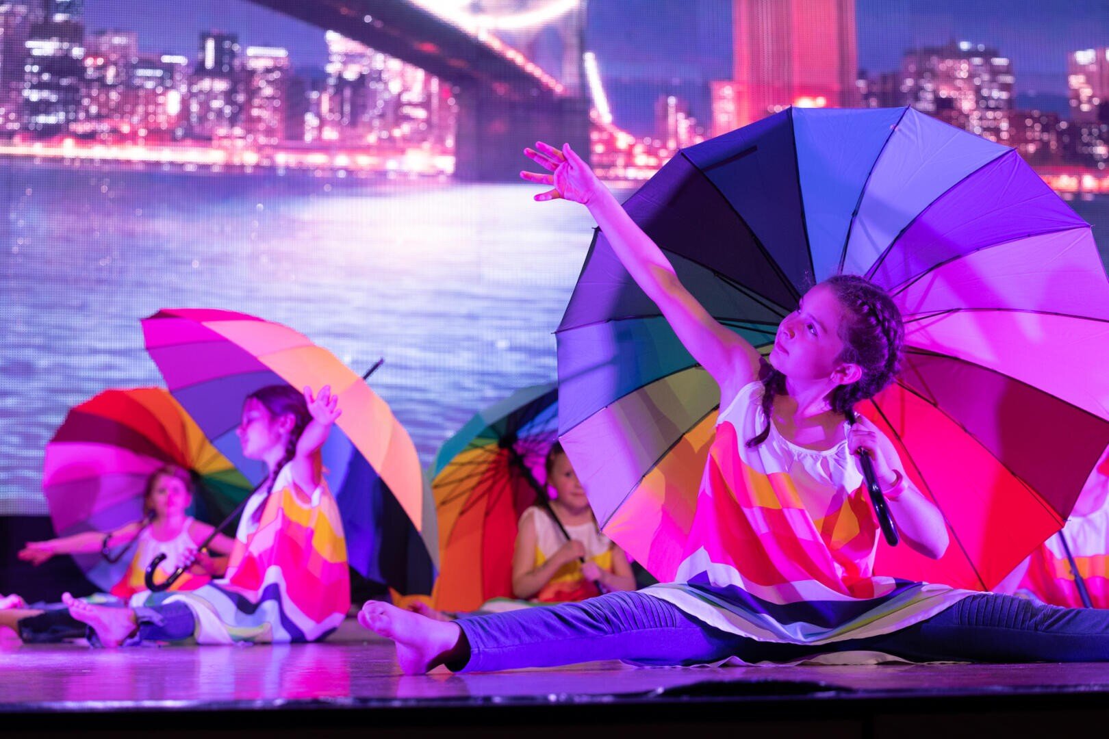 Junge Mädchen führen auf der Bühne einen Tanz auf, tragen bunte Outfits und halten große Regenbogenschirme. Der Hintergrund zeigt eine nächtliche Stadtsilhouette mit einer beleuchteten Brücke über dem Wasser. Ein Mädchen macht einen Spagat und streckt sich in die Höhe.