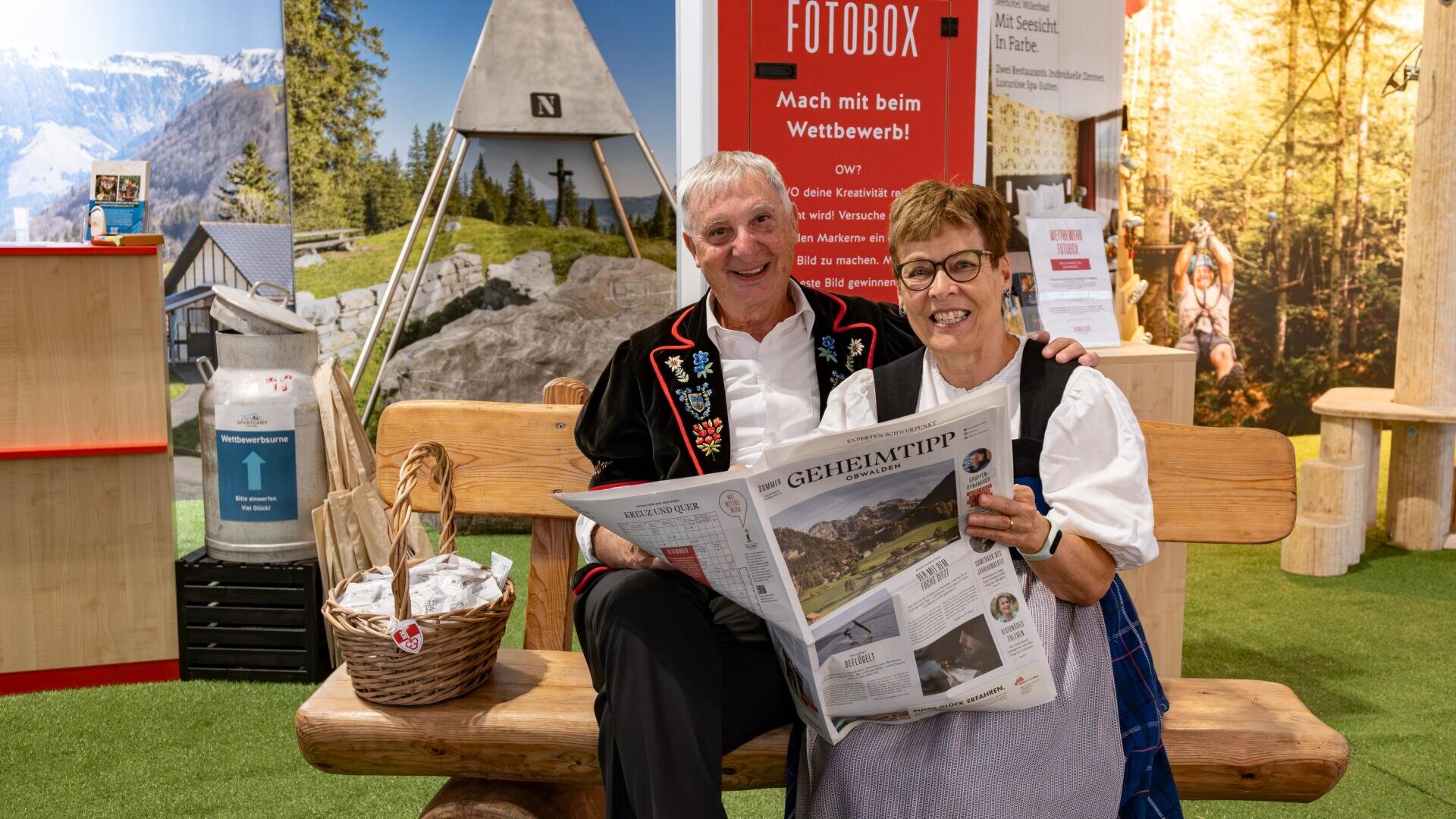 Ein älterer Mann und eine Frau in traditioneller Schweizer Kleidung sitzen auf einer Holzbank im Haus, lächeln und halten eine Zeitung in der Hand. Hinter ihnen stehen alpenländische Dekorationen, Poster und ein Korb mit Souvenirartikeln.