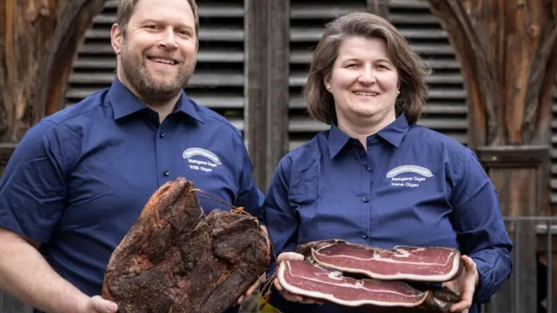 Zwei Personen in blauen Hemden, die große Stücke geräuchertes und gepökeltes Fleisch in den Händen halten, lächeln in die Kamera und stehen im Freien vor einem rustikalen Holzgebäude.