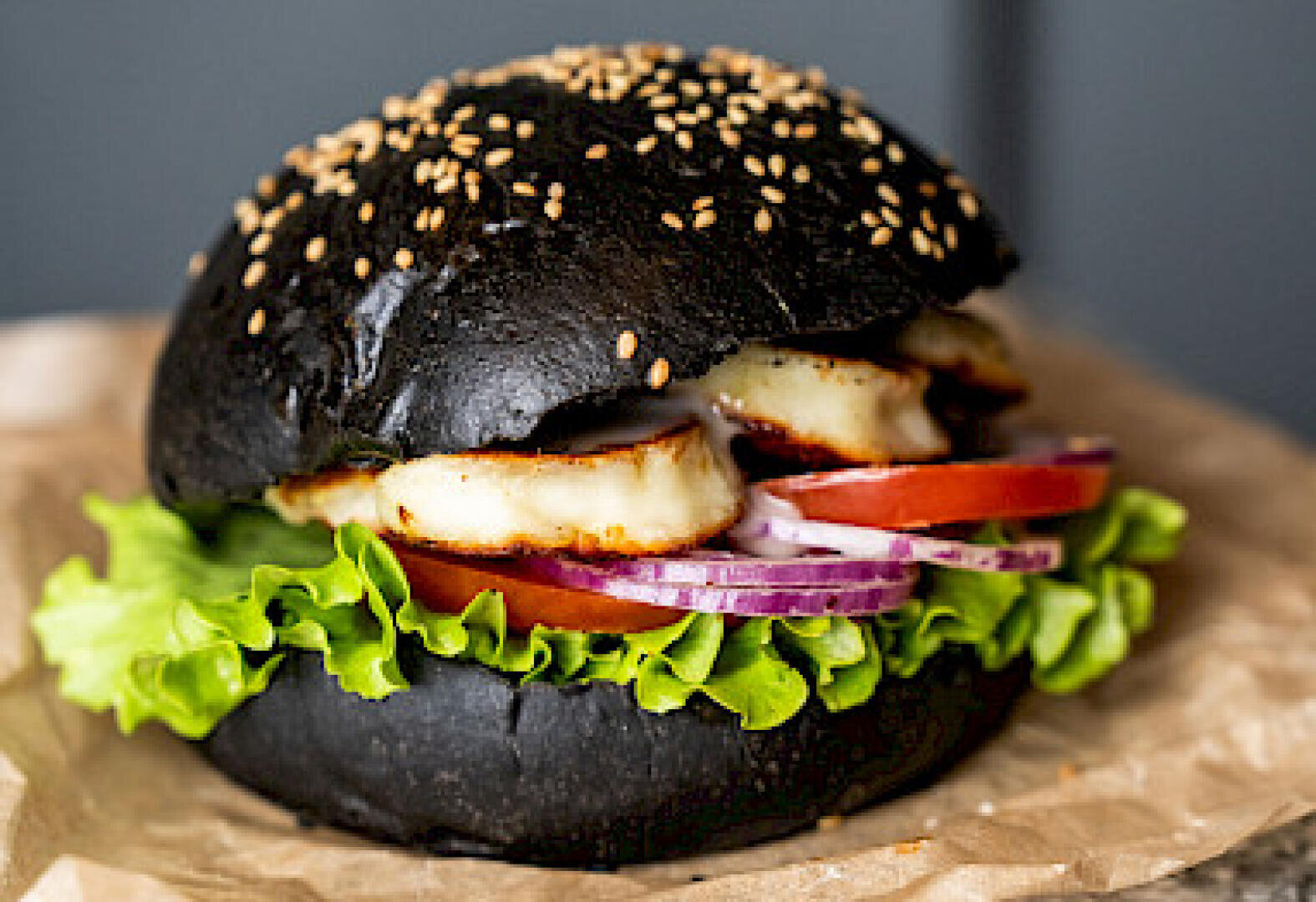 Ein schwarzer Sesambrötchen-Burger, gefüllt mit Salat, Tomaten, roten Zwiebelscheiben und gegrilltem Halloumi-Käse, auf braunem Pergamentpapier ruhend.