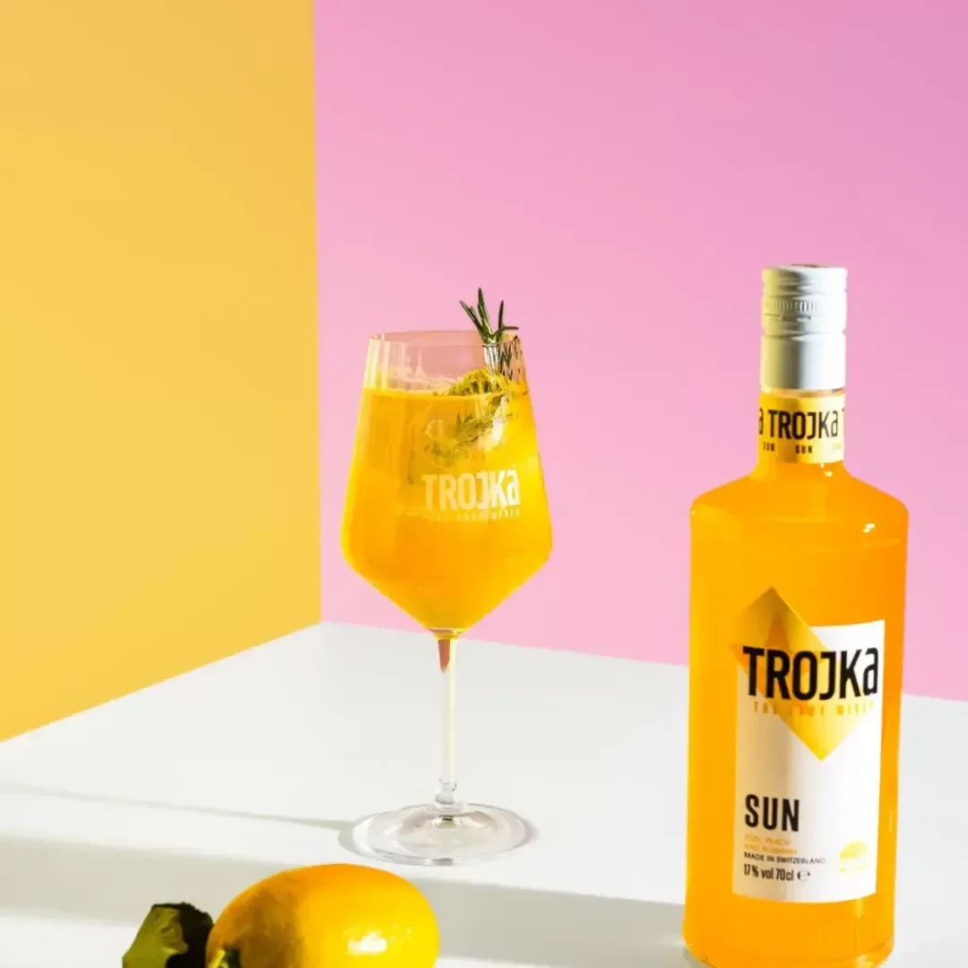 Eine gelbe Flasche Trojka-Sun-Likör steht neben einem Glas, das mit einem orange-gelben Getränk, Eis und einem Rosmarinzweig gefüllt ist. Eine Zitrone und grüne Blätter liegen in der Nähe; der Hintergrund ist gelb und rosa.