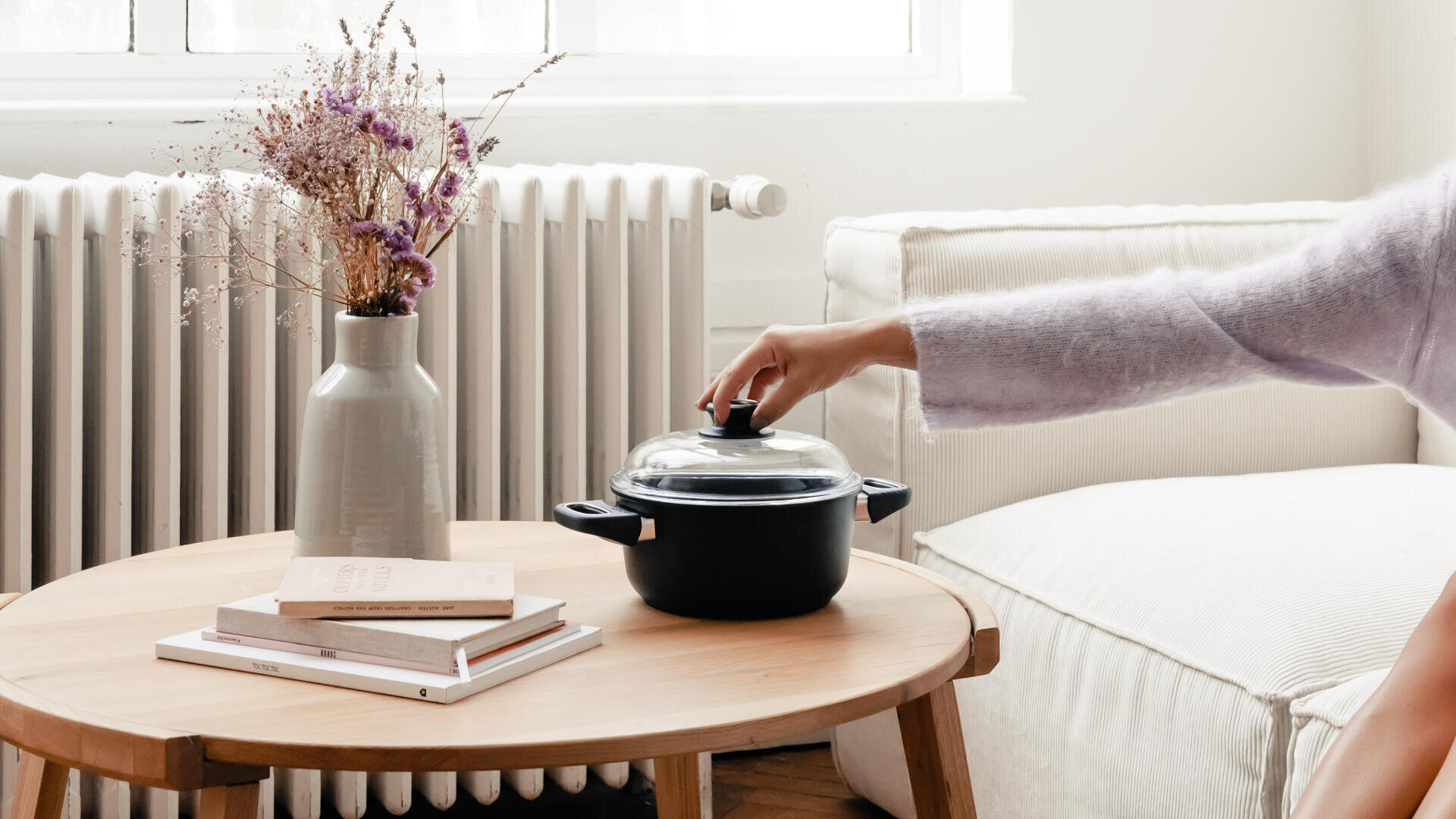 In einem hellen, gemütlichen Wohnzimmer hebt eine Person den Deckel eines kleinen schwarzen Topfes auf einem Holztisch neben einer Vase mit Trockenblumen und zwei geschlossenen Büchern ab.