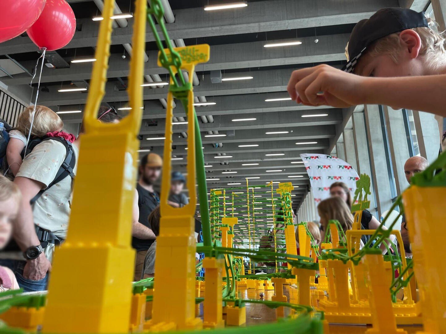 Ein Kind mit schwarzer Mütze spielt bei einer Indoor-Veranstaltung mit einer großen, bunten Spielzeug-Achterbahn. Im Hintergrund sind andere Menschen und rote Luftballons zu sehen, über denen moderne Deckenleuchten angebracht sind.