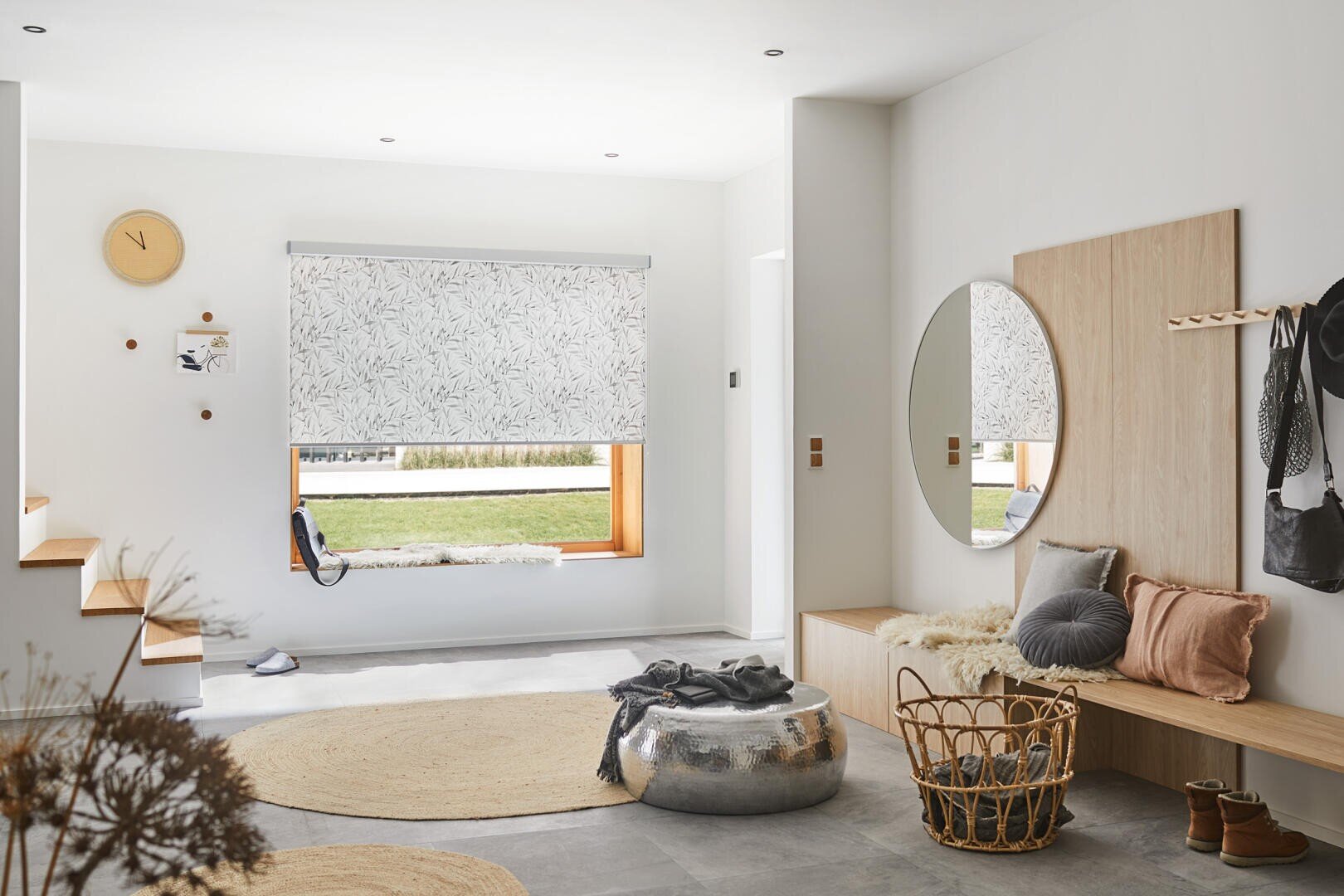 Ein moderner Eingangsbereich in neutralen Tönen, ein runder Spiegel über einer Holzbank mit Kissen, ein metallischer Hocker, ein geflochtener Korb, ein beiger Teppich, Wandhaken mit Hängeartikeln und ein großes Fenster mit gemusterten Jalousien.