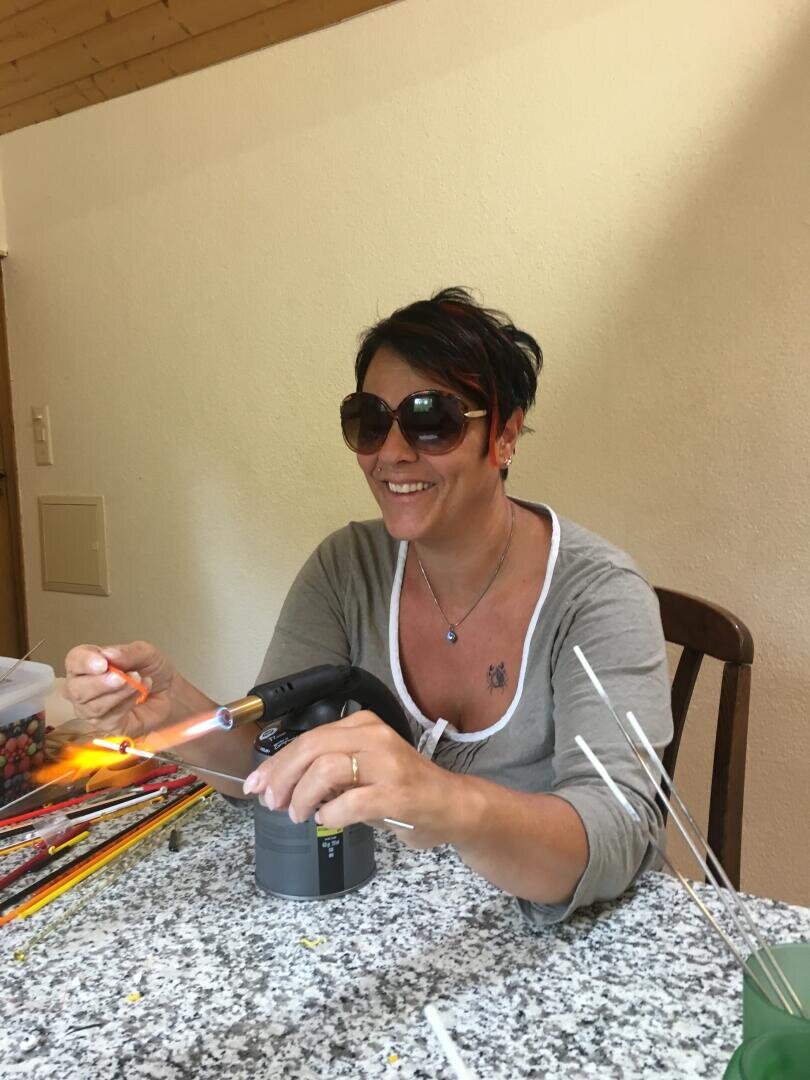 Eine Frau mit Sonnenbrille und grauem Oberteil lächelt, während sie mit einer Fackel an einem gesprenkelten Tisch mit Glasstäben arbeitet.