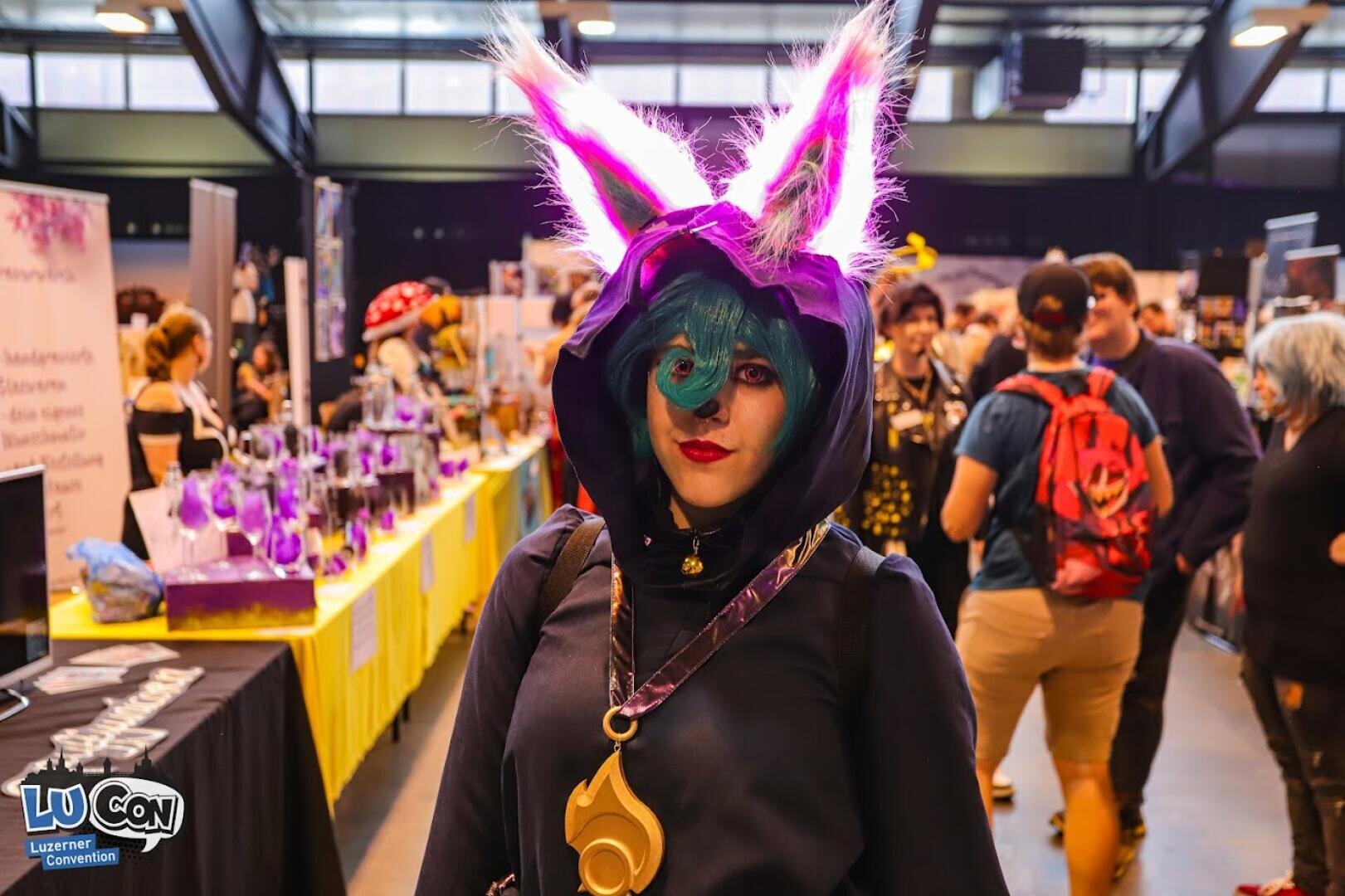 Eine Person in einem farbenfrohen Fantasy-Cosplay mit großen, pelzigen lila Ohren und einer blauen Perücke steht in einer belebten Kongresshalle, umgeben von Verkäufertischen und anderen Teilnehmern. Im Hintergrund sind Banner von Cosplay-Veranstaltungen zu sehen.