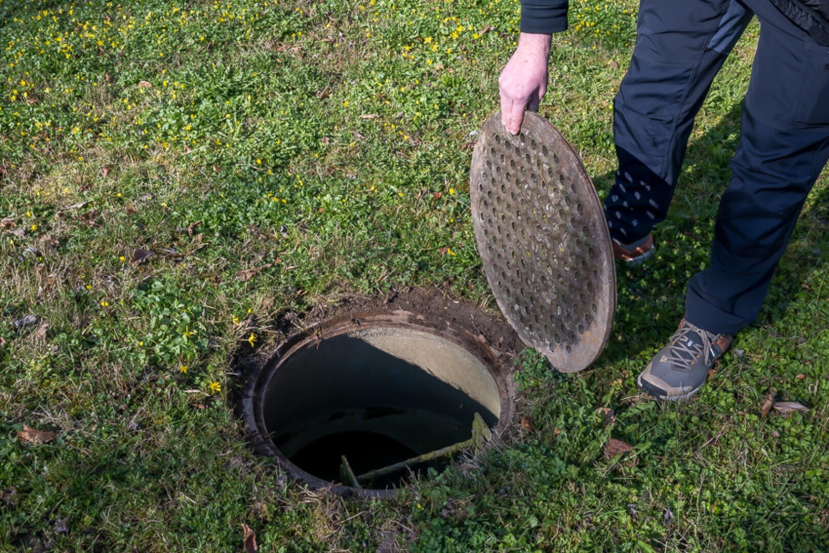Eine Person hebt einen runden Gullydeckel aus einer kreisrunden Öffnung im Boden, umgeben von grünem Gras und gelben Blumen.
