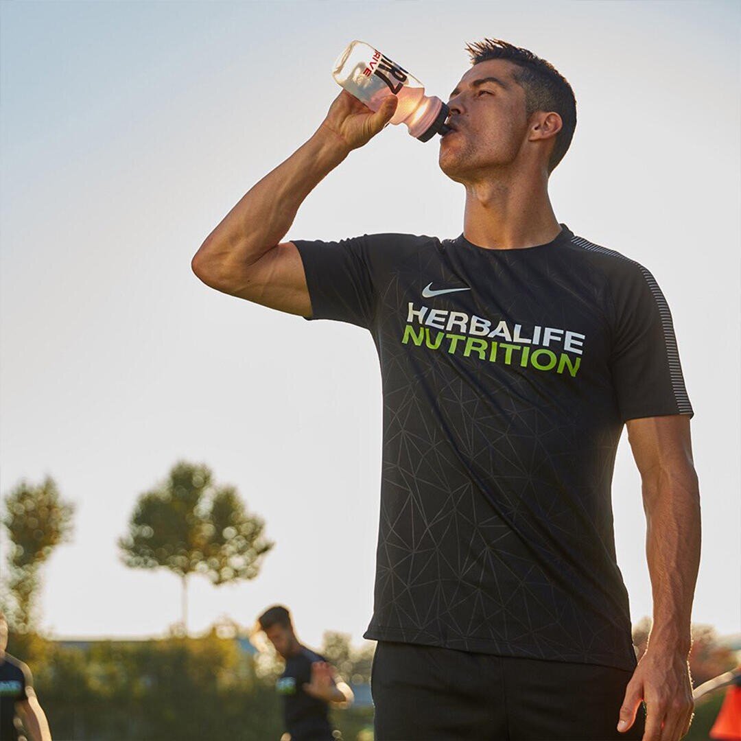 Ein Mann, der ein schwarzes Herbalife Nutrition T-Shirt trägt, trinkt im Freien aus einer Wasserflasche, während im Hintergrund Bäume und Sonnenlicht zu sehen sind.