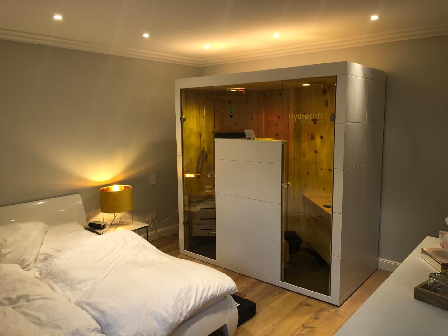 Ein modernes Schlafzimmer mit einem ordentlich gemachten Bett, einer gelb leuchtenden Lampe auf dem Nachttisch und einer großen weißen Hydrosoft-Saunakabine mit Glastüren in der Ecke des Raumes.