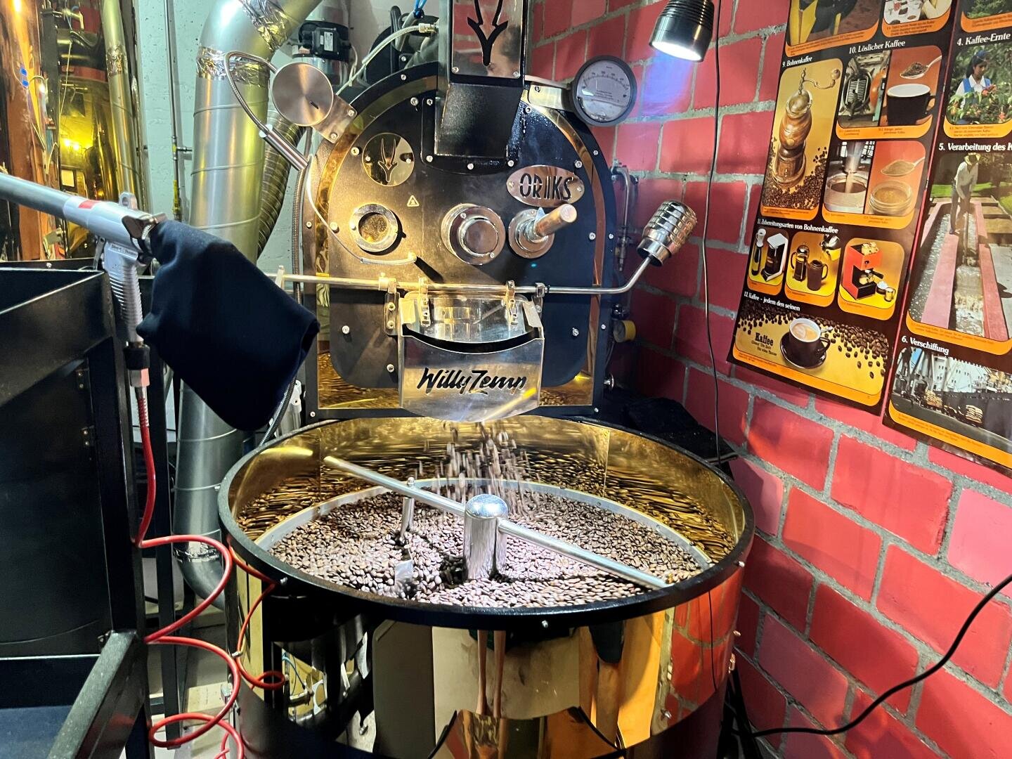 Eine glänzende Kaffeeröstmaschine, in der Kaffeebohnen geröstet werden, neben einer Backsteinmauer und Plakaten zum Thema Kaffee, mit Metallrohren, Lampen und verschiedenen Geräten drum herum.
