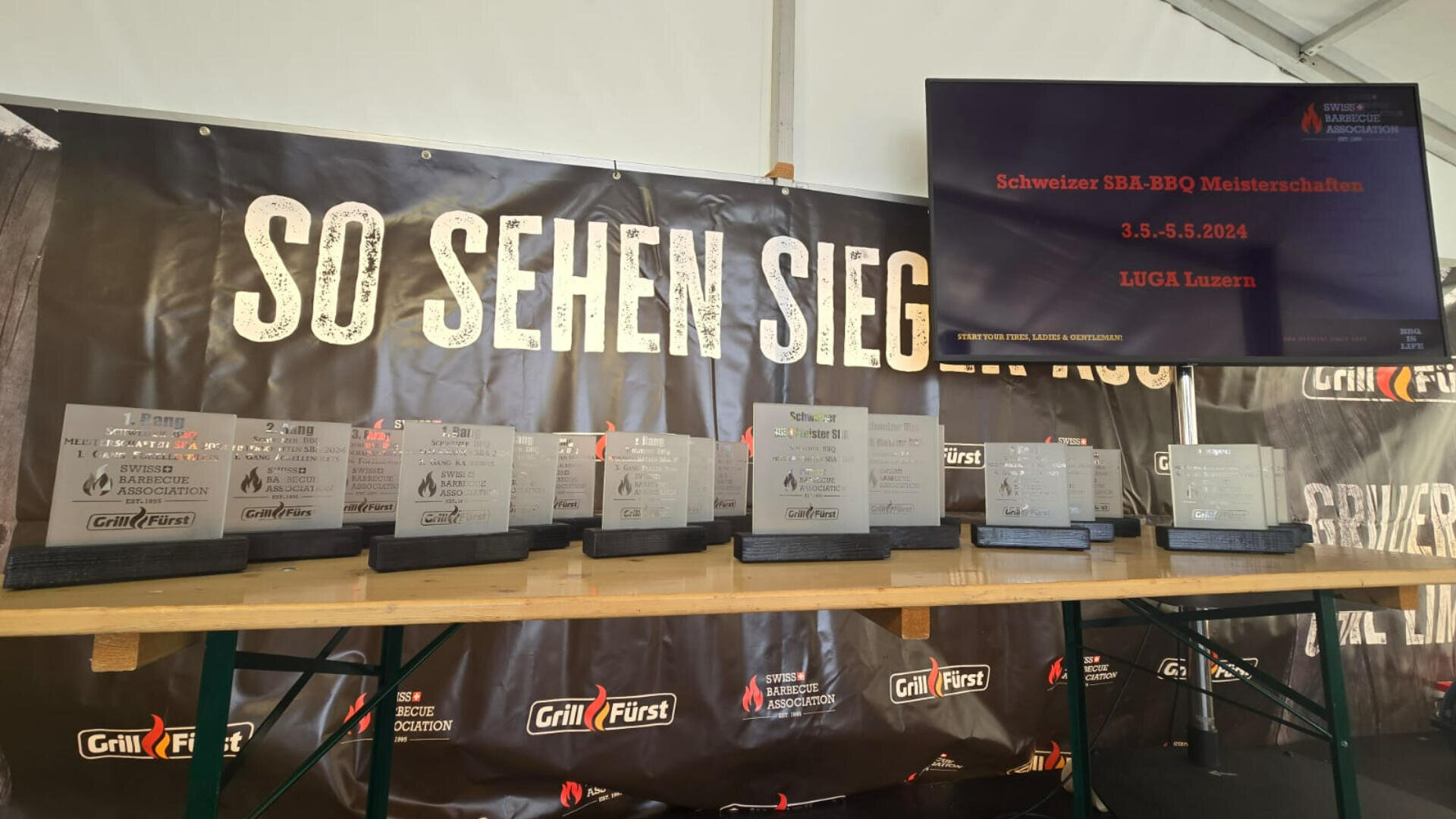 Eine Reihe von metallenen BBQ-Wettbewerbstrophäen auf einem Tisch vor einem dunklen Banner mit der Aufschrift SO SEHEN SIEGER AUS. Ein Bildschirm zeigt Veranstaltungsinformationen an: Schweizer SBL-BBQ Meisterschaften, 3.-5.5.2024, LUGA Luzern.