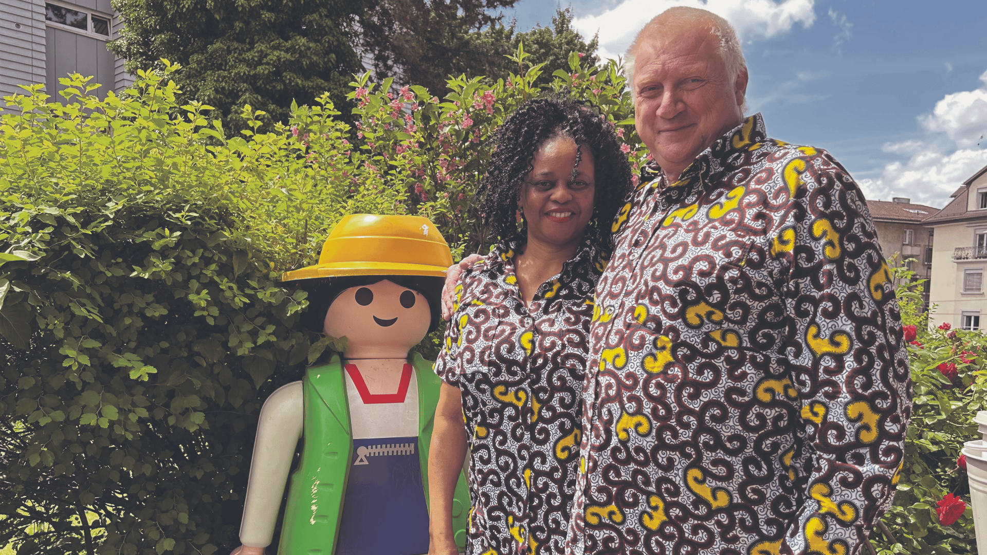 Ein lächelndes Paar in passenden gemusterten Hemden steht im Freien neben einer großen Puppe im Playmobil-Stil mit gelbem Hut, grüner Weste und rotem Overall. Im Hintergrund sind üppiges Grün und ein blauer Himmel mit Wolken zu sehen.