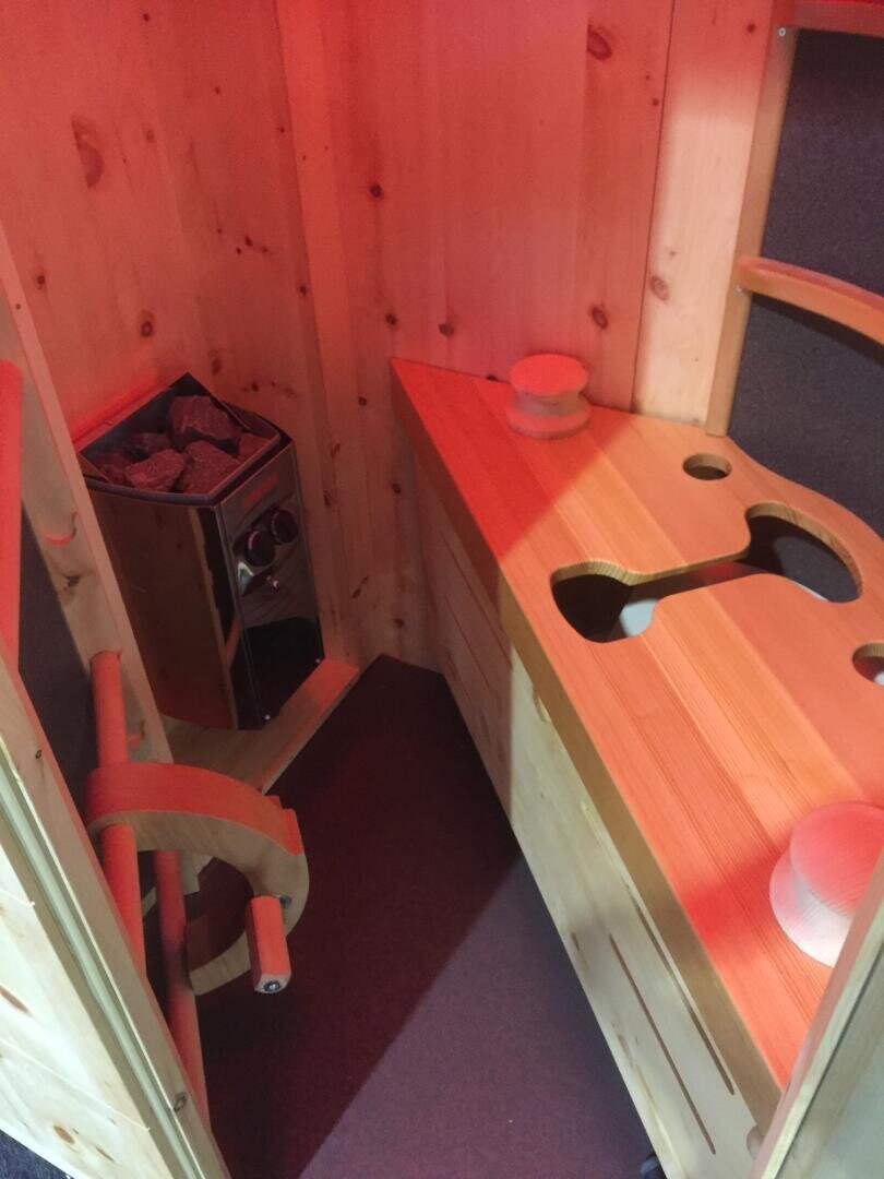 Ein kleiner hölzerner Saunaraum mit einem mit Steinen gefüllten Ofen, Bänken und einem hölzernen Eimer auf der Theke. Die Wände und das Innere sind aus hellem Holz, und die Beleuchtung verleiht dem Raum einen warmen, rötlichen Schimmer.
