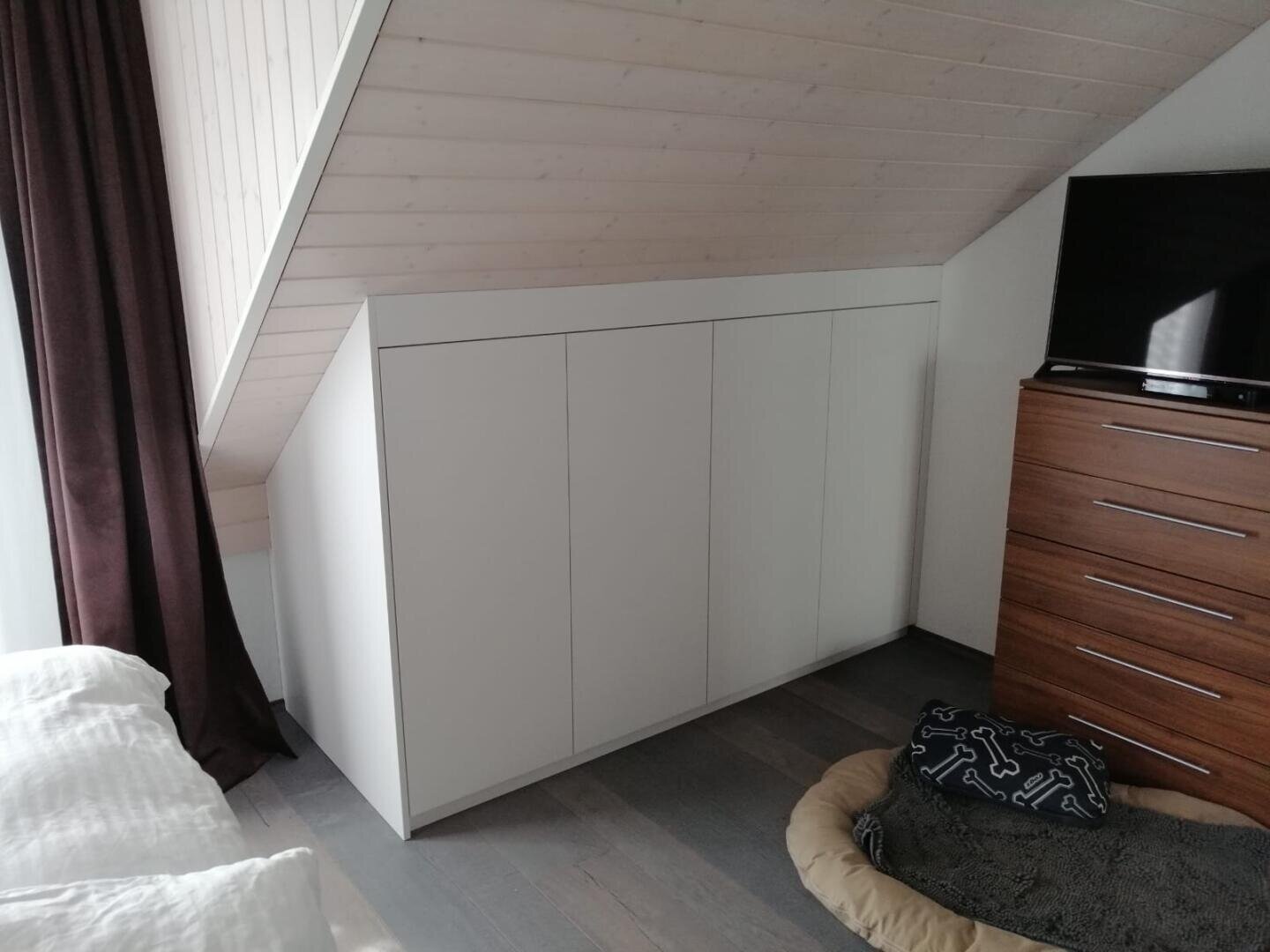 Ein weißer Einbauschrank passt unter eine schräge Holzdecke in einem Schlafzimmer. In der Nähe stehen eine hölzerne Kommode mit einem Fernseher darauf, ein großes Hundebett und ein Teil eines Bettes mit weißem Bettzeug.