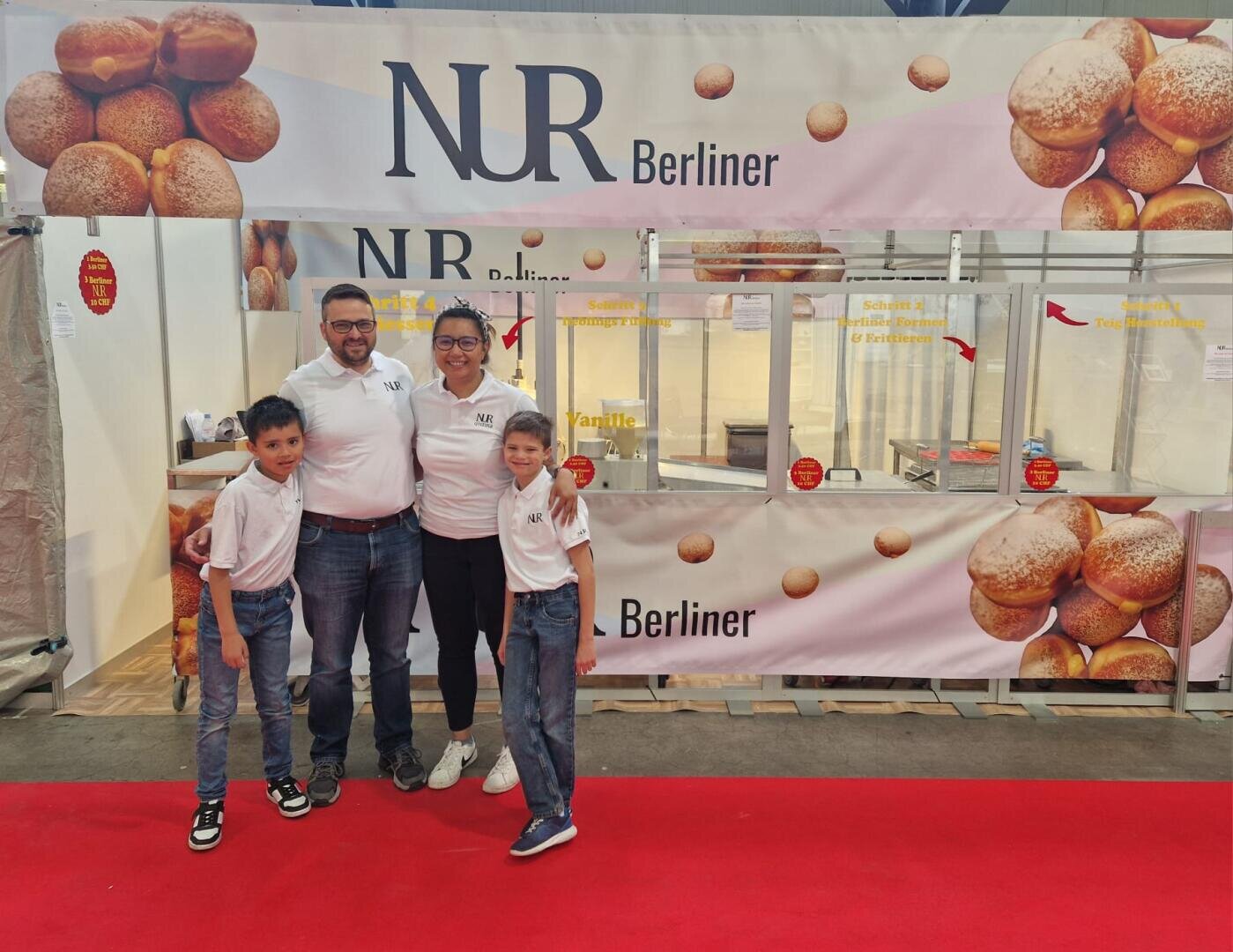 Eine vierköpfige Familie, gekleidet in passende weiße Hemden und Jeans, steht lächelnd vor einem Bäckereistand namens NR Berliner, der mit Bildern von runden Backwaren dekoriert ist. Vor dem Stand liegt ein roter Teppich.
