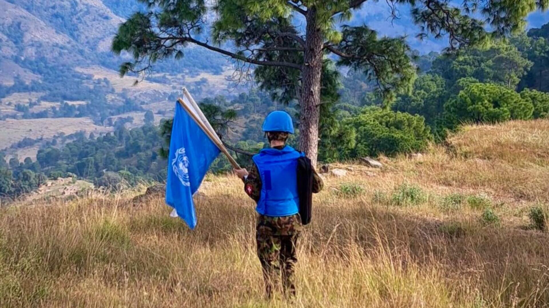 Eine Person in Militärtarnung und mit blauem Helm steht auf einem grasbewachsenen Gelände und hält eine Flagge der Vereinten Nationen in der Nähe eines Baumes, während im Hintergrund Berge und Grünflächen zu sehen sind.