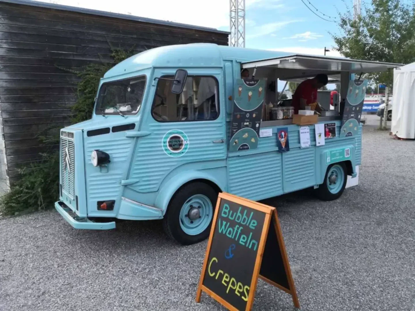 Ein alter blauer Imbisswagen parkt mit geöffnetem Seitenfenster auf einem Schotterplatz und bedient Kunden. Ein Kreideschild an der Vorderseite wirbt für Bubble Waffles und Crêpes. Der Truck hat Retro-Details und bunte Dekorationen.