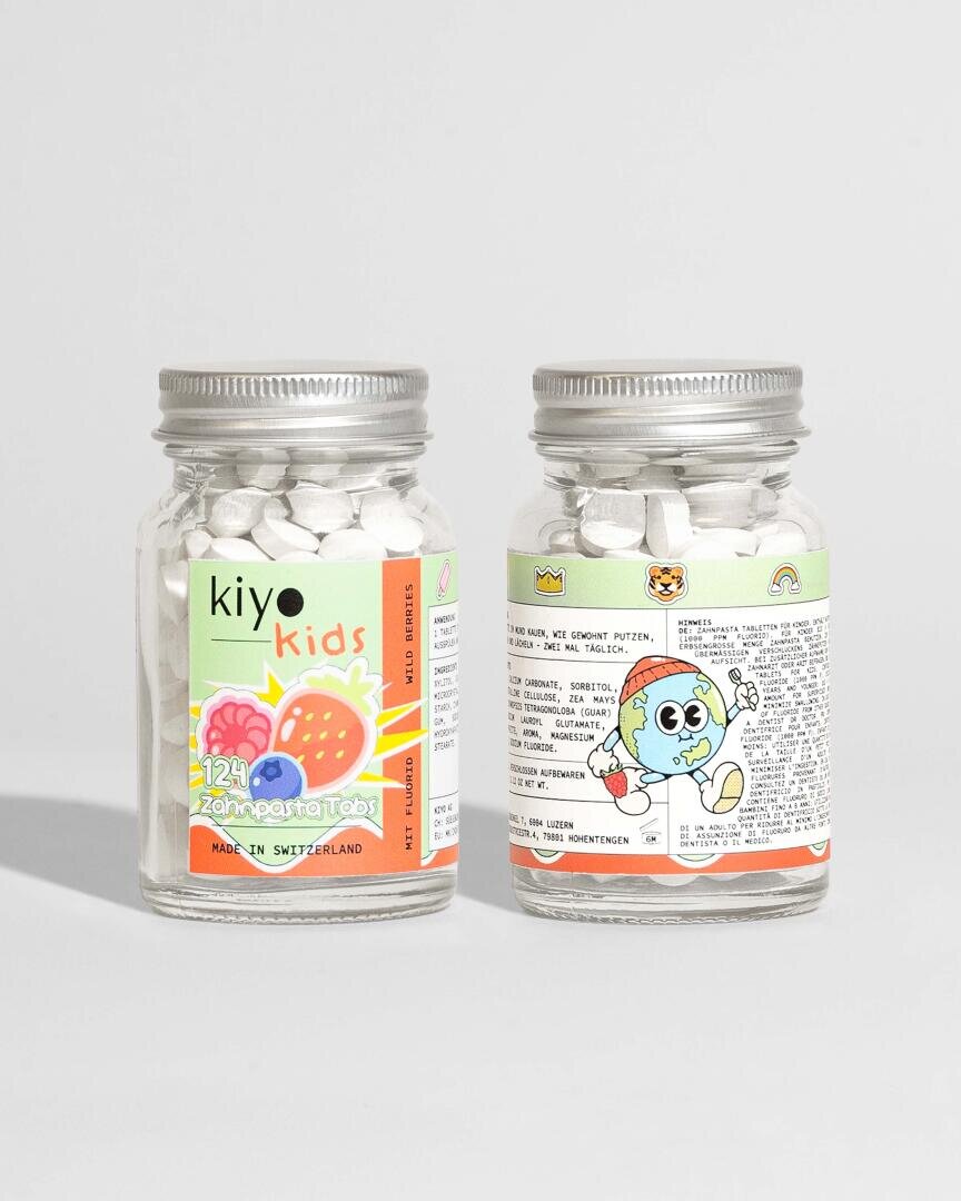 Zwei kleine Gläser mit Kiyo Kids Zahnpastatabletten mit silbernen Deckeln. Das Etikett auf der Vorderseite ist farbenfroh mit einem Regenbogenzahnmotiv, während die Rückseite eine Zeichentrickfigur und Produktinformationen in deutscher Sprache zeigt.