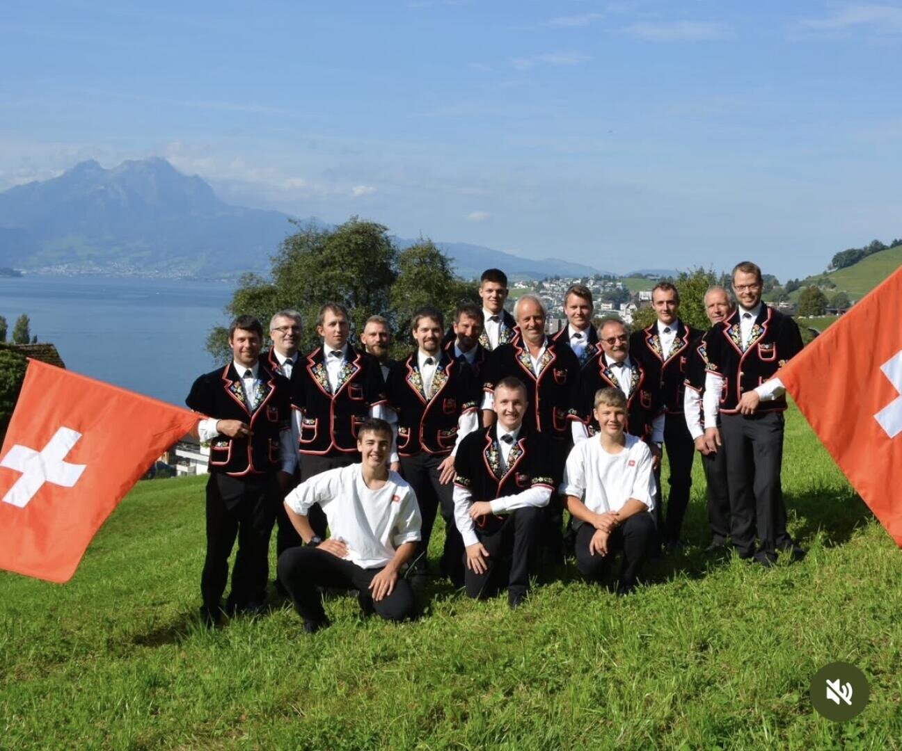Eine Gruppe von Männern, einige in traditioneller Schweizer Tracht und andere in weißen Hemden, posieren im Freien auf einem grasbewachsenen Hügel mit Schweizer Fahnen. An einem klaren Tag sind im Hintergrund ein See, Berge und Bäume zu sehen.