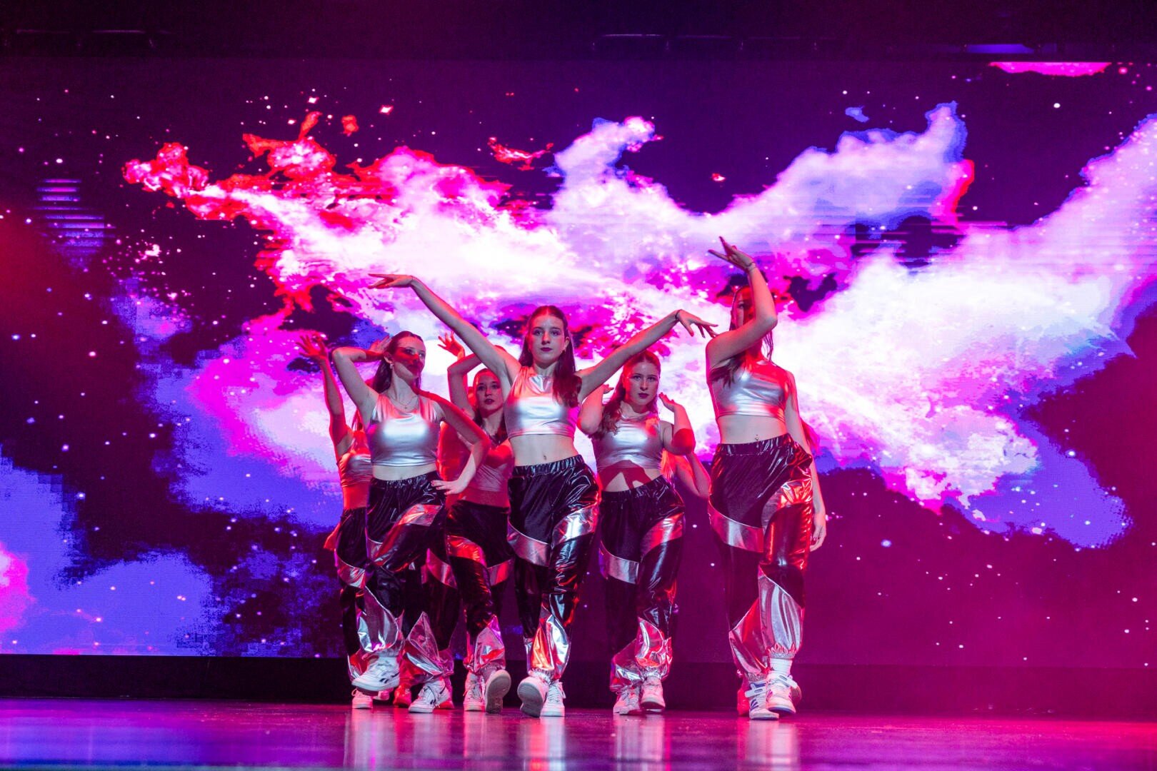 Eine Gruppe von Tänzerinnen und Tänzern in silbern glänzenden Kostümen tanzt energisch auf der Bühne vor einem lebhaften, farbenfrohen kosmischen Hintergrund mit rosa, lila und weißen wolkenähnlichen Mustern.
