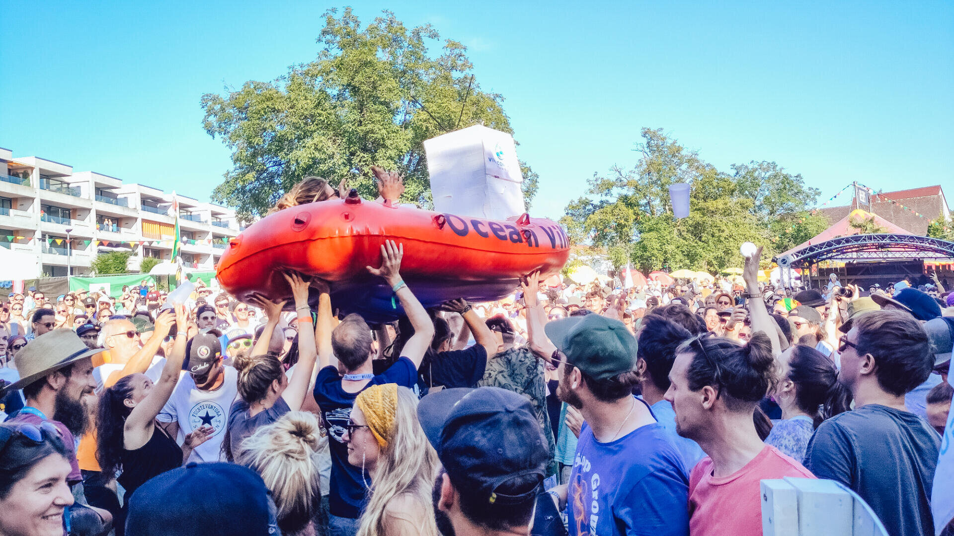 Eine große Menschenmenge auf einem Festival im Freien hebt eine Person in einem aufblasbaren orangefarbenen Boot über ihre Köpfe. Die Menschen lächeln, tragen Sommerkleidung und Hüte, im Hintergrund sind Bäume und Gebäude zu sehen.